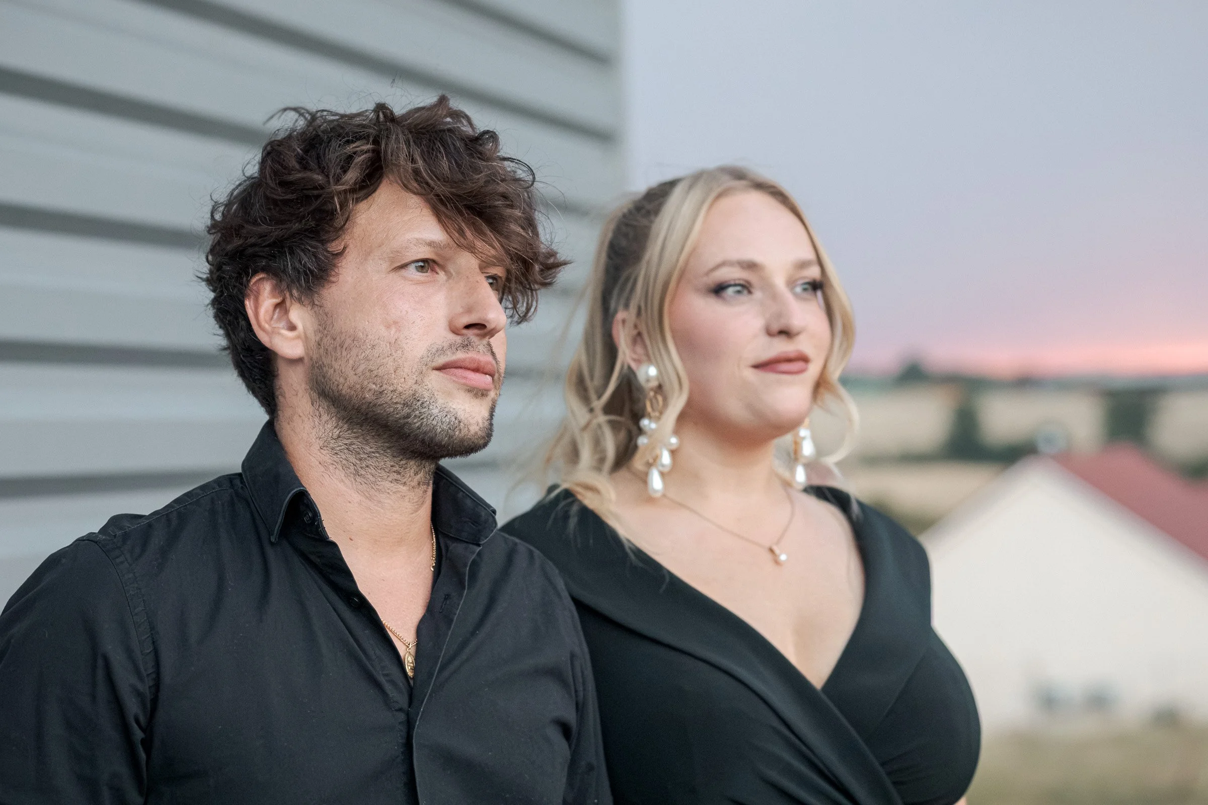 Deux personnes regardent au loin, un homme avec des cheveux bouclés foncés et une femme aux cheveux blonds, portant des boucles d'oreilles pendantes, tous deux vêtus de noir, avec un fond de ciel au crépuscule.