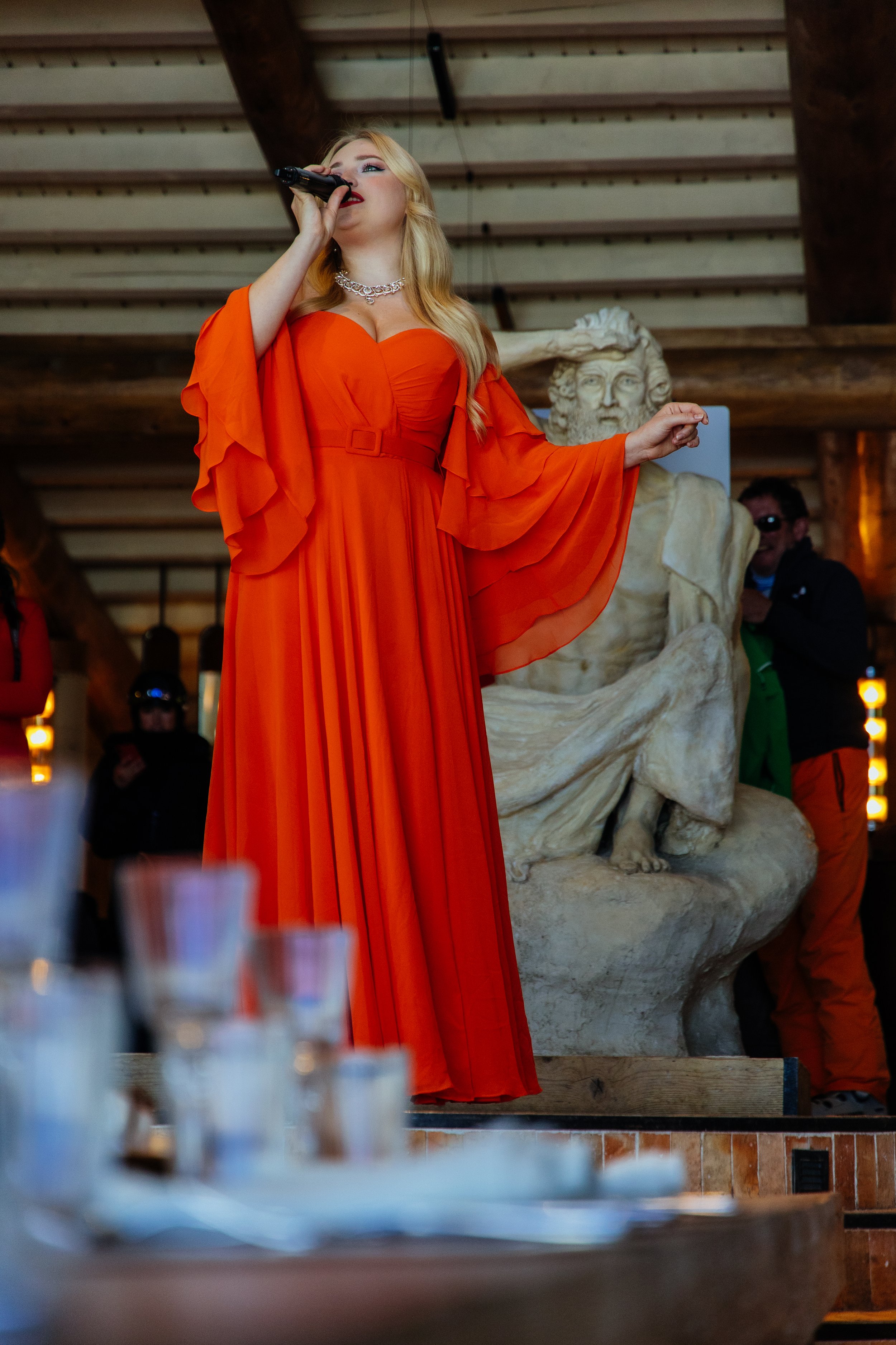 Une femme en robe orange chantant avec un microphone lors d'une performance, avec une sculpture en marbre en arrière-plan.