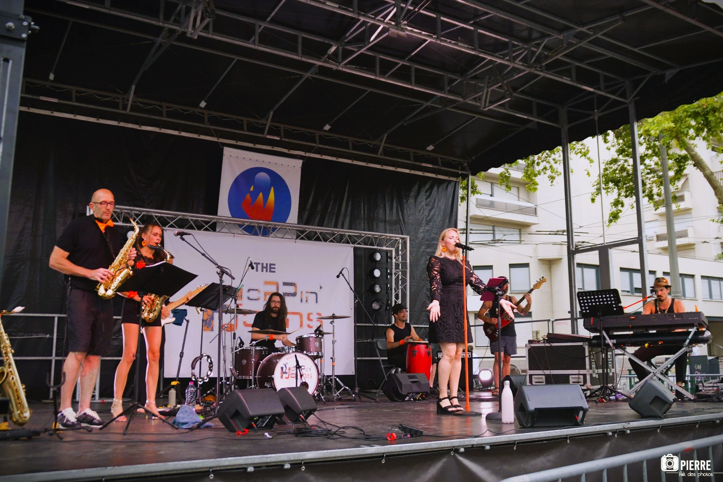 Groupe de musiciens sur une scène extérieure lors d'un concert, avec une femme au chant et une autre à la guitare, en arrière-plan un batteur et un percussionniste, devant un panneau promotionnel.