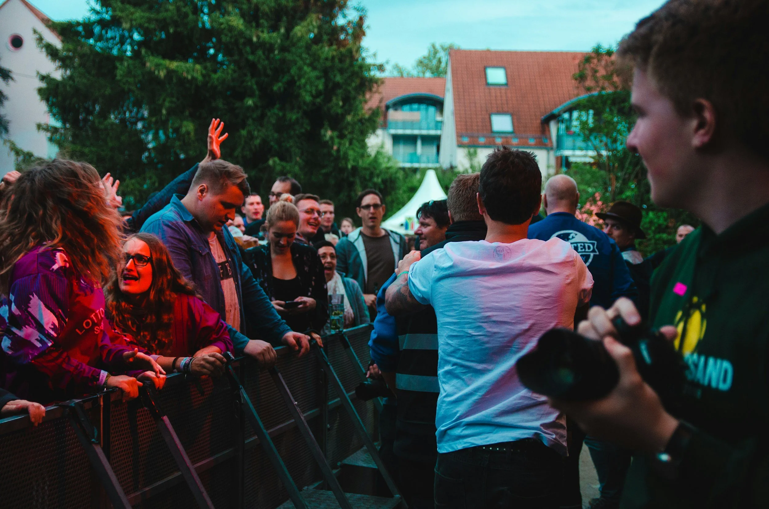 A crowd of people gathered behind a metal barricade, engaging in conversation and raising their hands, with a backdrop of trees, houses with red roofs, and tents, during an outdoor event.