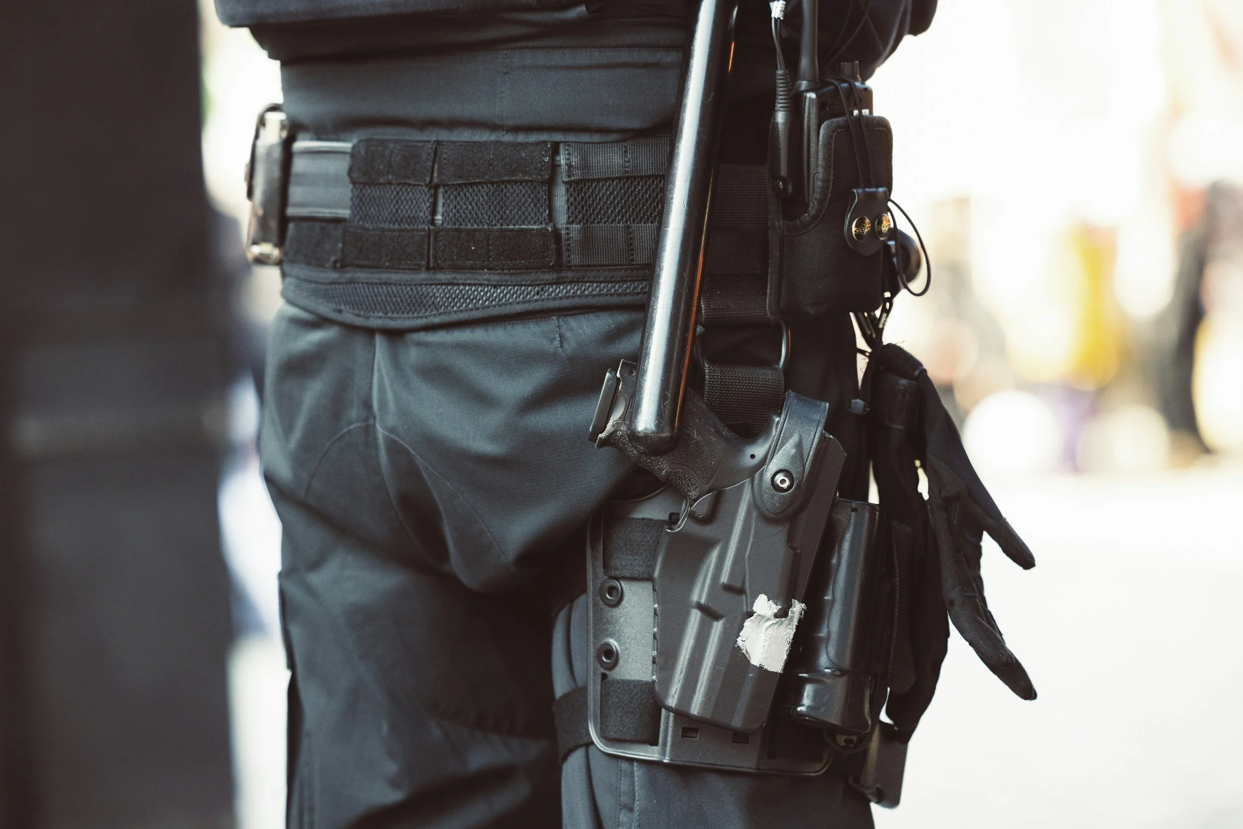 Close-up of a security officer's tactical belt with a holstered firearm, handcuffs, and communication device, worn over dark tactical pants.