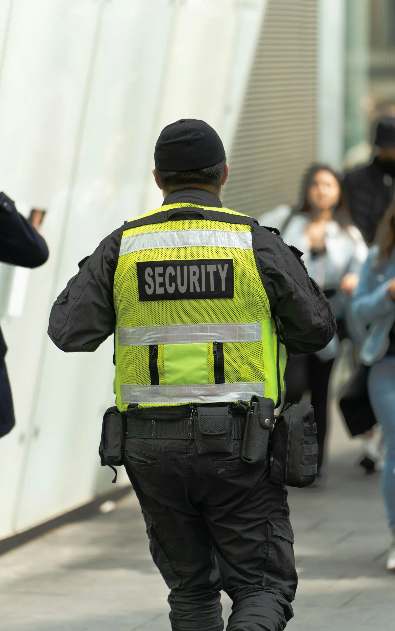 A security officer wearing a black uniform and a high-visibility yellow vest with 'SECURITY' written on the back, walking in an indoor public space with blurred people in the background.