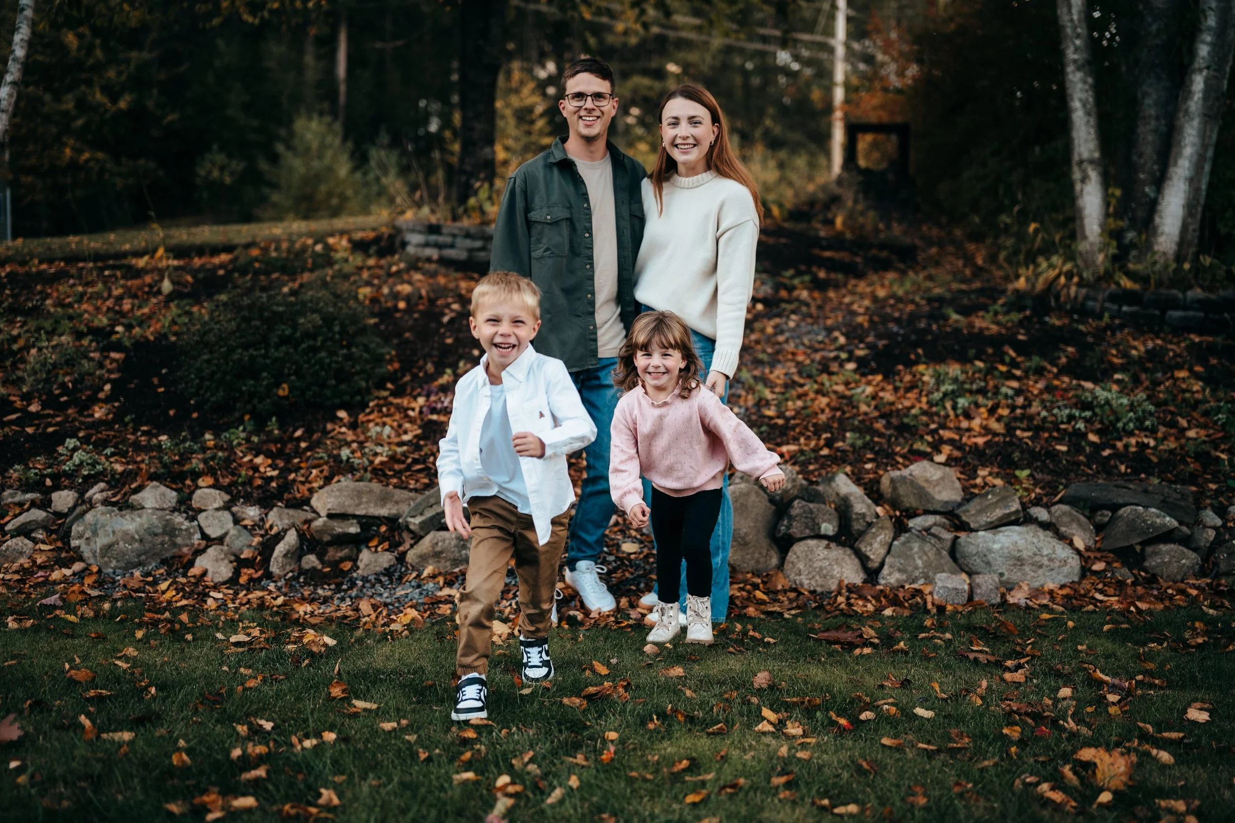 A smiling family of four running outdoors among fall leaves, with a man, woman, and two children, in a wooded backyard with fallen leaves and rocks.