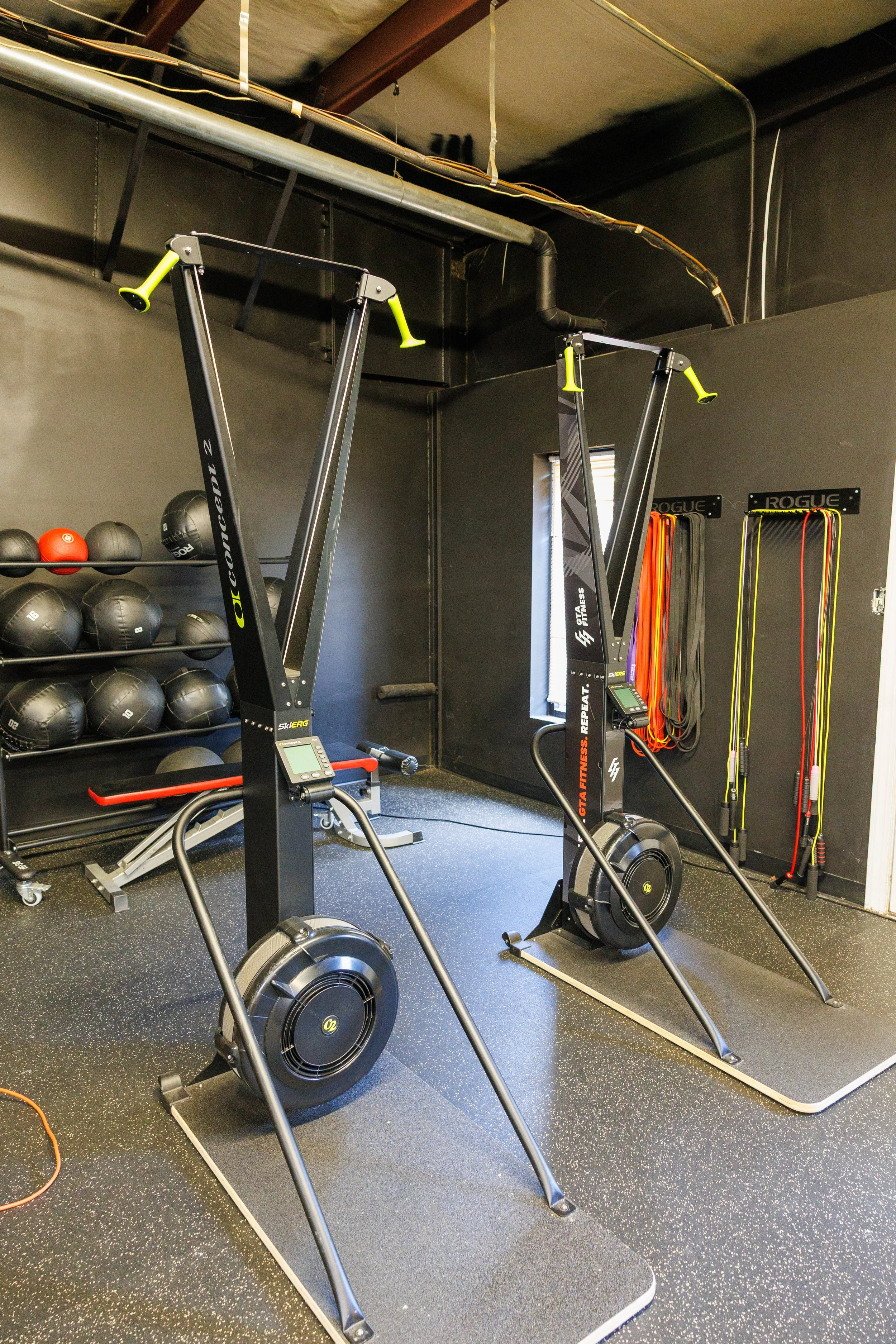Two concept2 rowing machines in a gym with black walls, orange resistance bands, and a shelf of black medicine balls.
