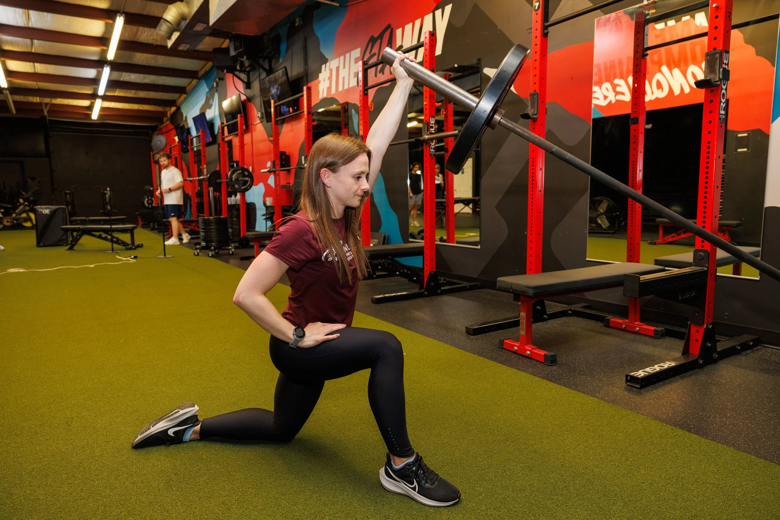 A woman in a gym performing a lunge exercise while holding a barbell overhead.