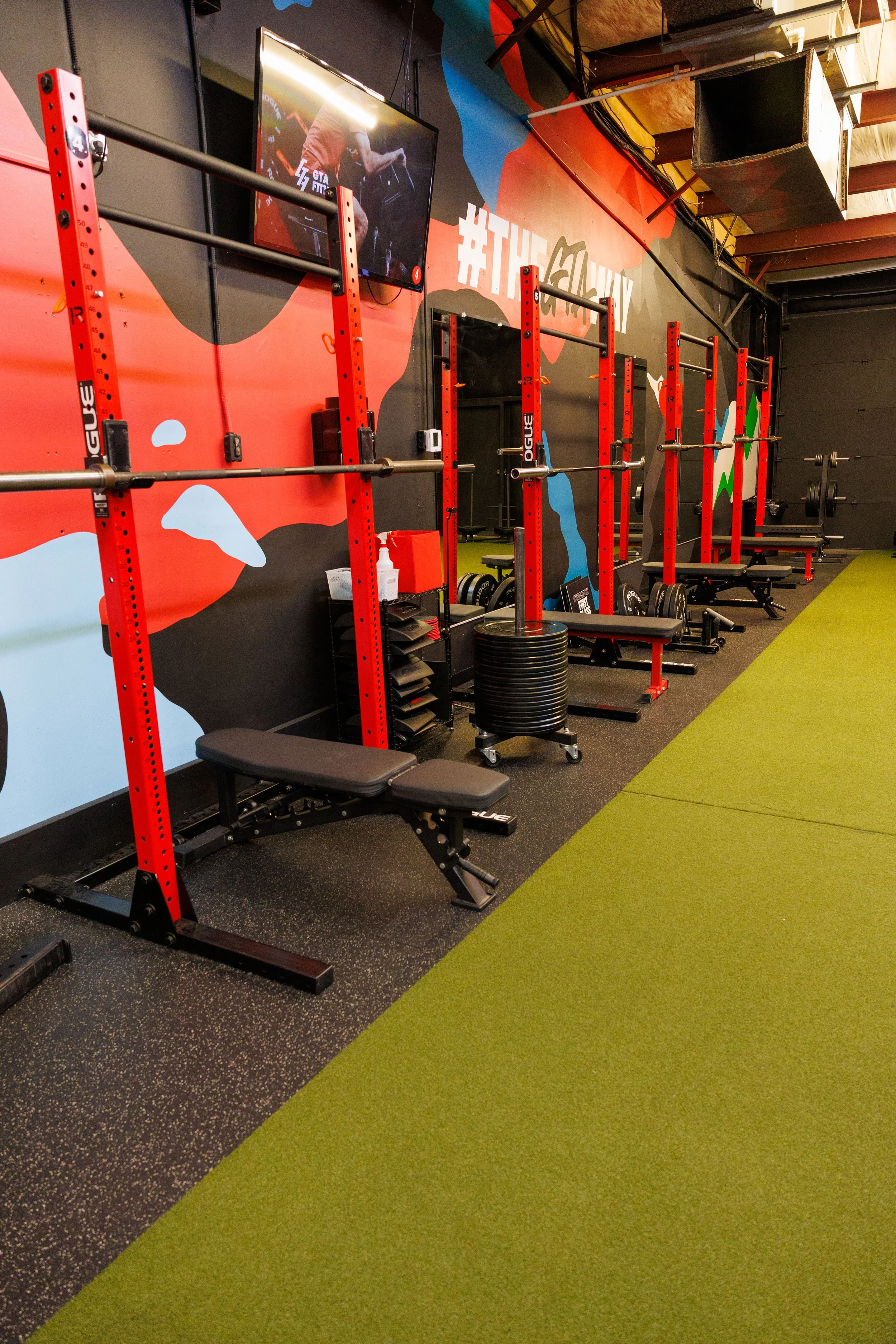Empty gym workout area with red squat racks, benches, weight plates, and a green and black flooring, with a mural and TV screen on the wall.