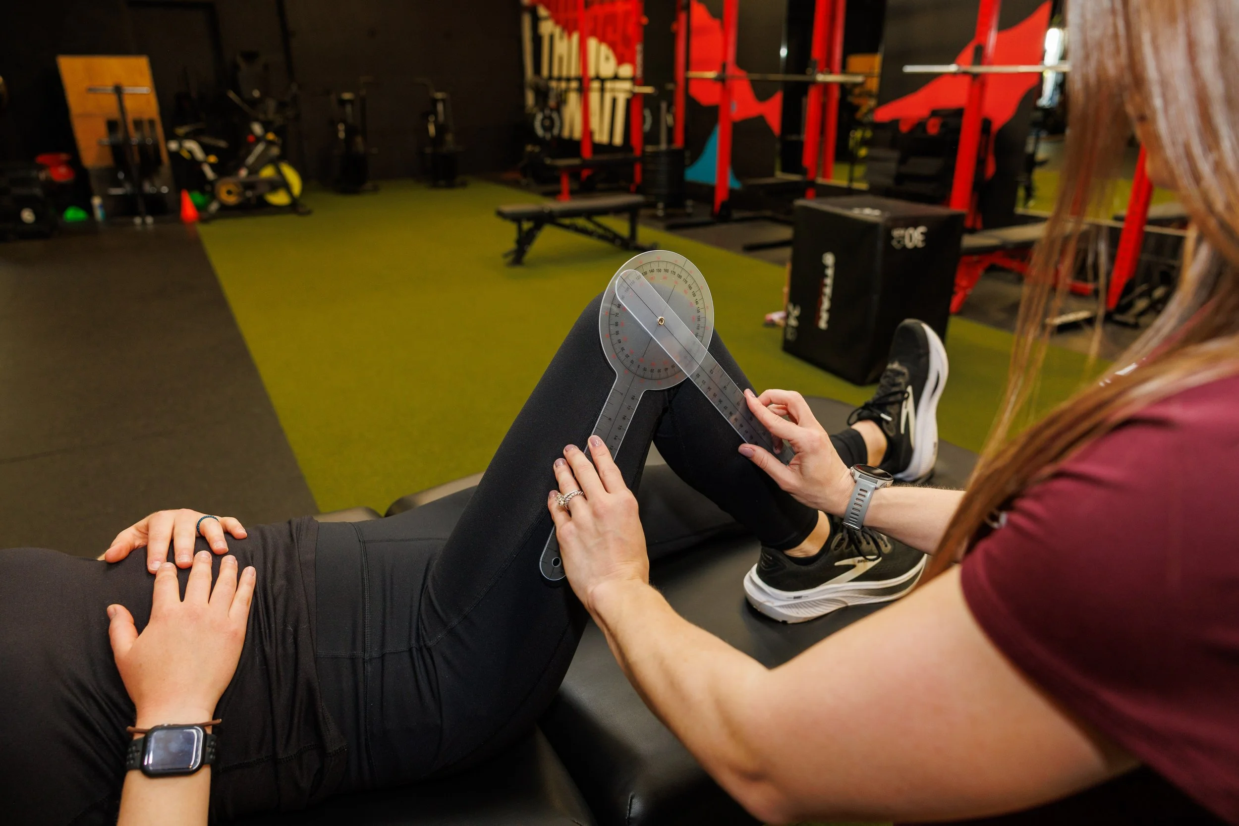 A woman is conducting a physical therapy assessment on a person lying down on a medical table in a gym, using a specialized goniometer to measure the knee joint's angle.