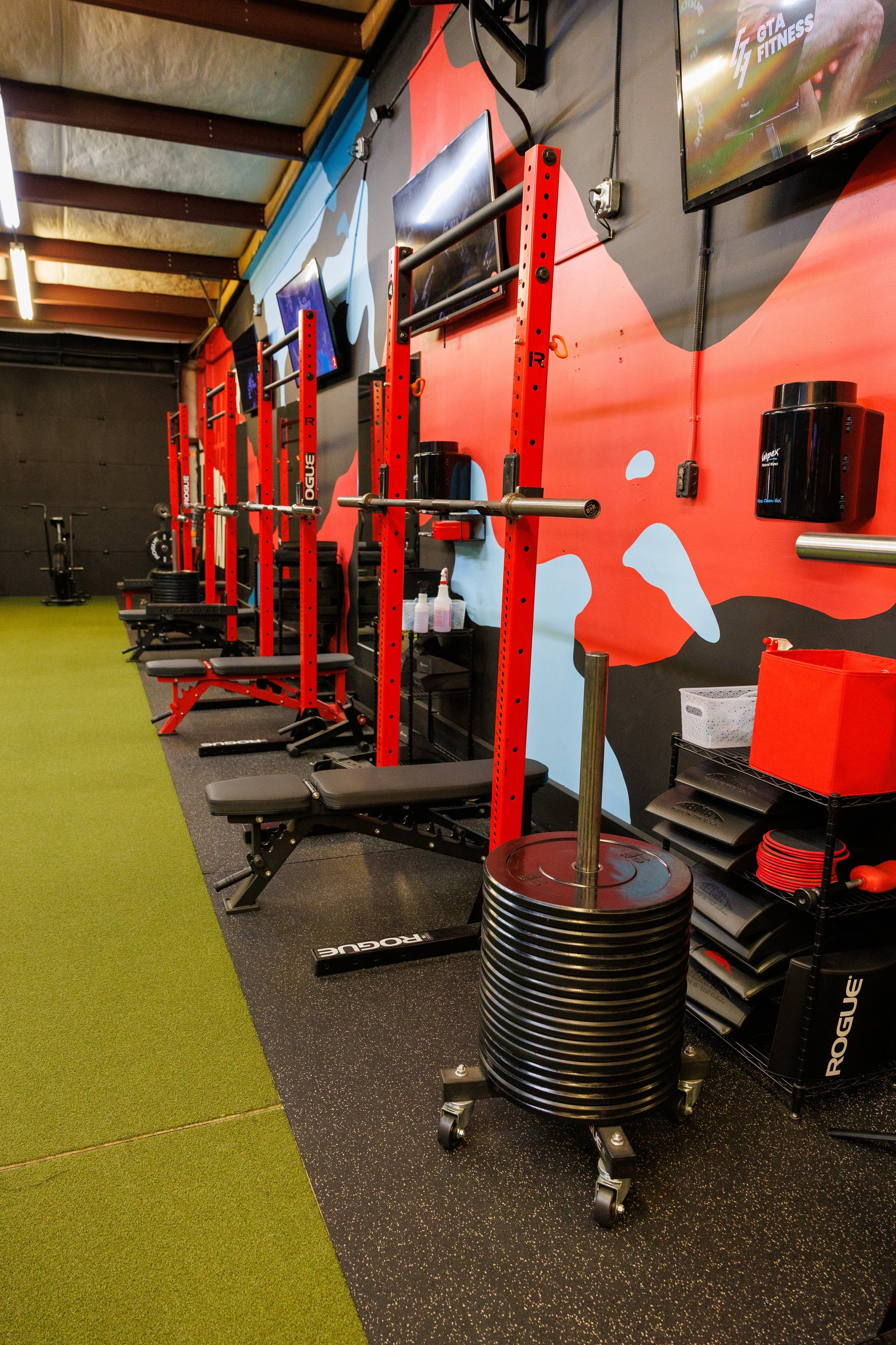 An interior view of a gym with red power racks, benches, weight plates on a cart, and wall-mounted TVs.