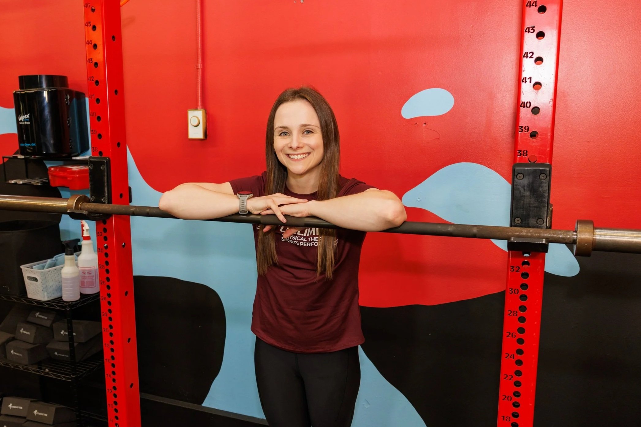 A woman smiling and leaning on a barbell in a gym with a red and blue wall behind her.