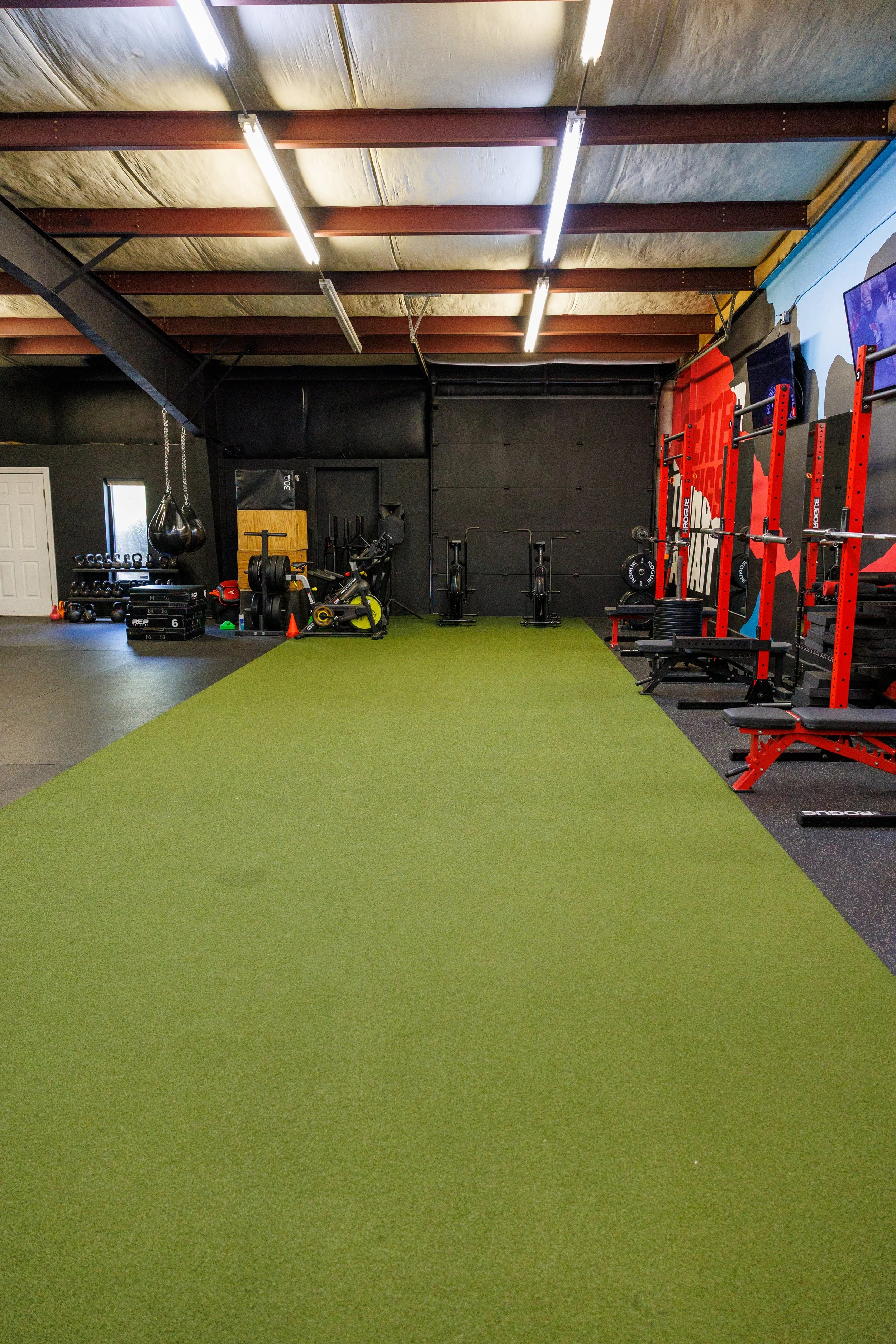 Inside view of a gym with green turf, red squat racks, weight plates, kettlebells, and fitness equipment.