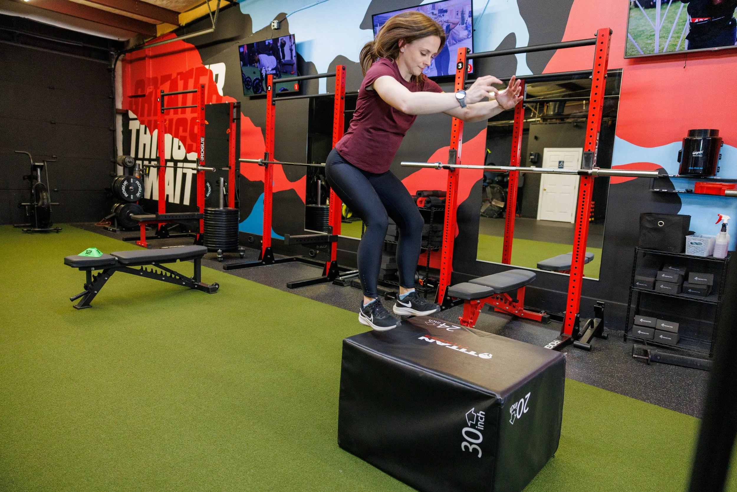 A woman jumping onto a black padded box during a workout in a gym with red and black equipment and a green carpeted floor.