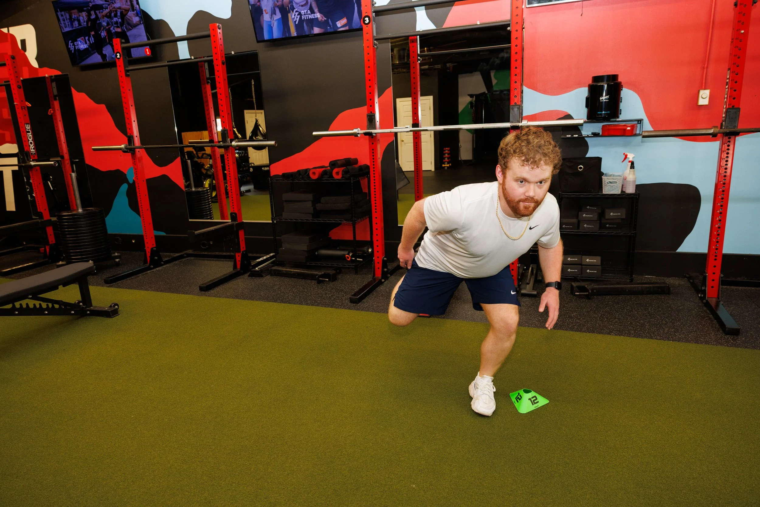 A man performing a workout exercise in a gym, placing his right hand on a green marker on the floor, wearing a white t-shirt, navy shorts, and white sneakers, with gym equipment and colorful wall art in the background.