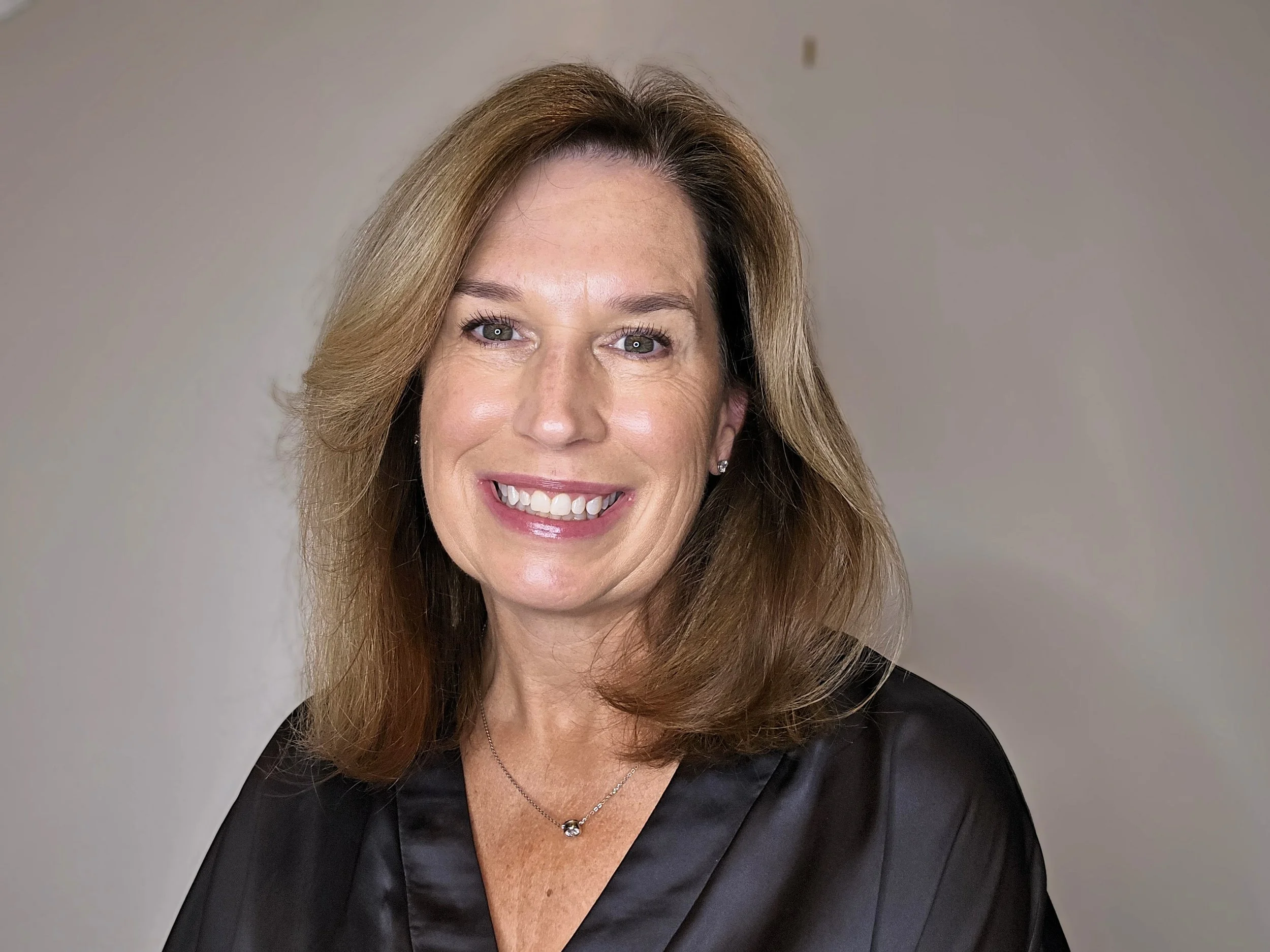 A professional woman with shoulder-length light brown hair, wearing a black satin blouse and a delicate necklace, smiling against a plain background.