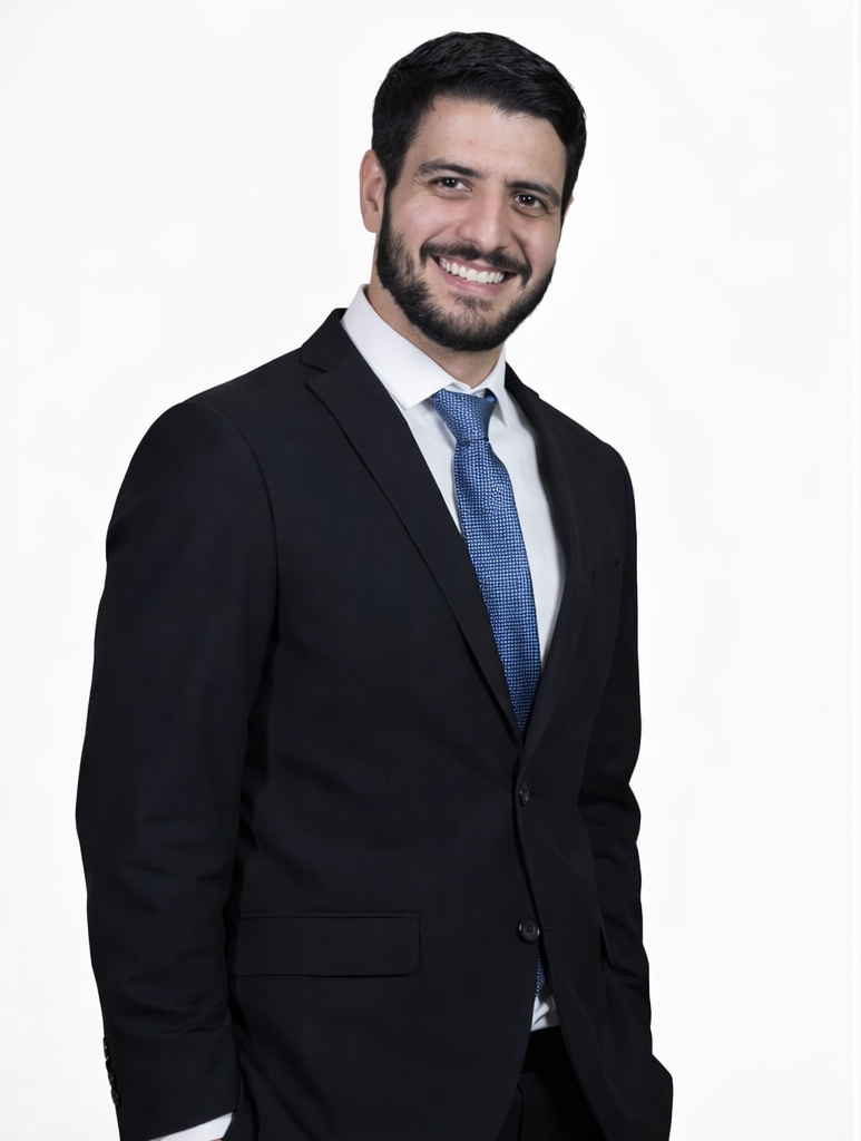 Homem de terno preto, camisa branca e gravata azul, sorrindo, em fundo branco.
