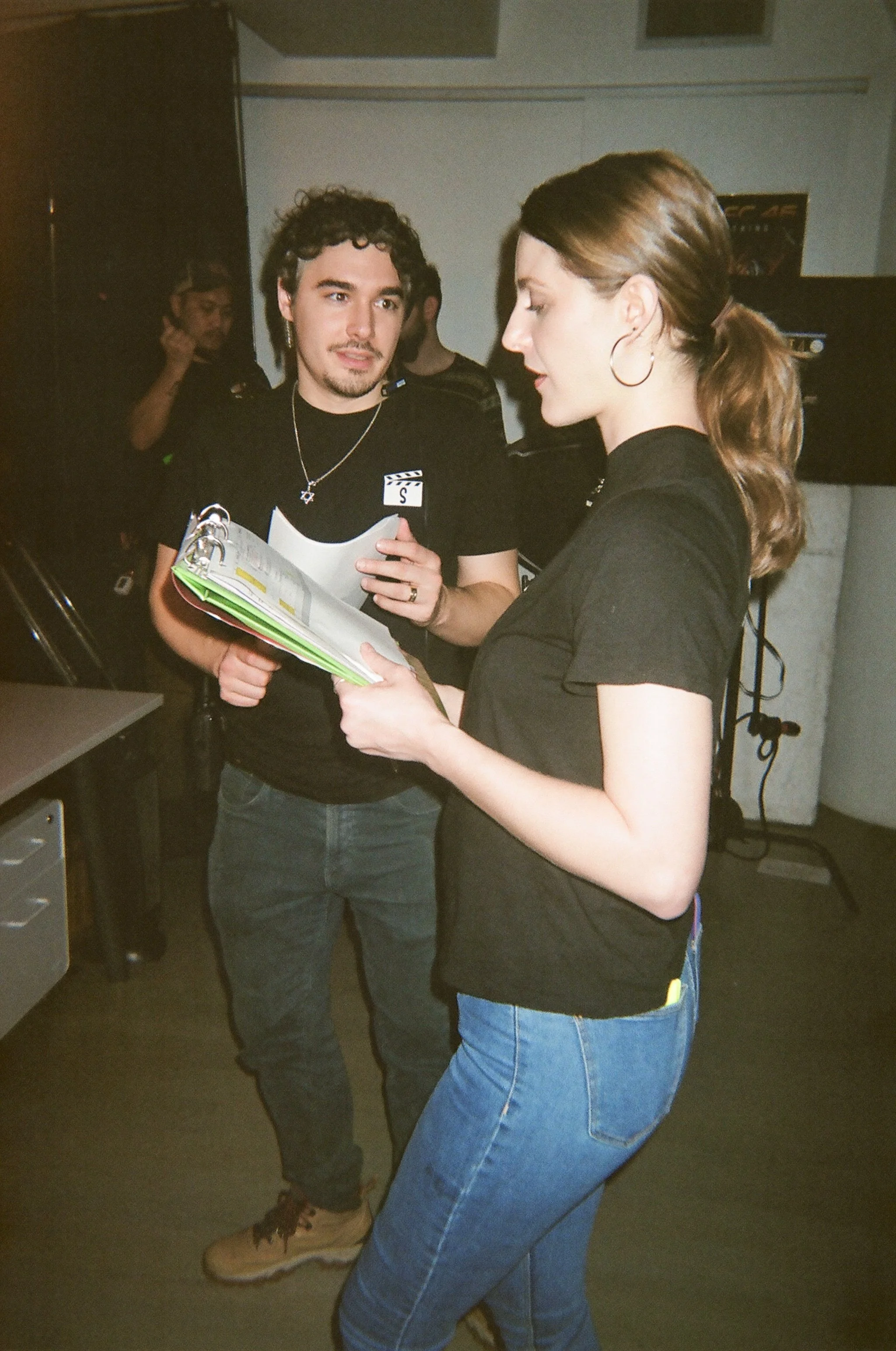 Two people, a man and a woman, are looking at a script or a book together indoors. The woman is wearing a black shirt and blue jeans, and the man is wearing a black T-shirt and dark jeans. There are other people and filming equipment in the backgroun
