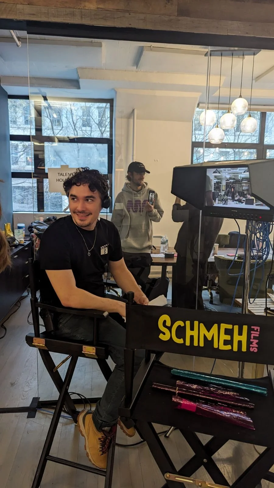 A young man with curly hair, wearing a black t-shirt and headphones, smiling and sitting on a director's chair. Behind him, another person is taking a photo with a smartphone. The setting is indoors with large windows and round hanging lights. There are film rolls on a table in the foreground, and a large monitor displaying a reflection of the scene.