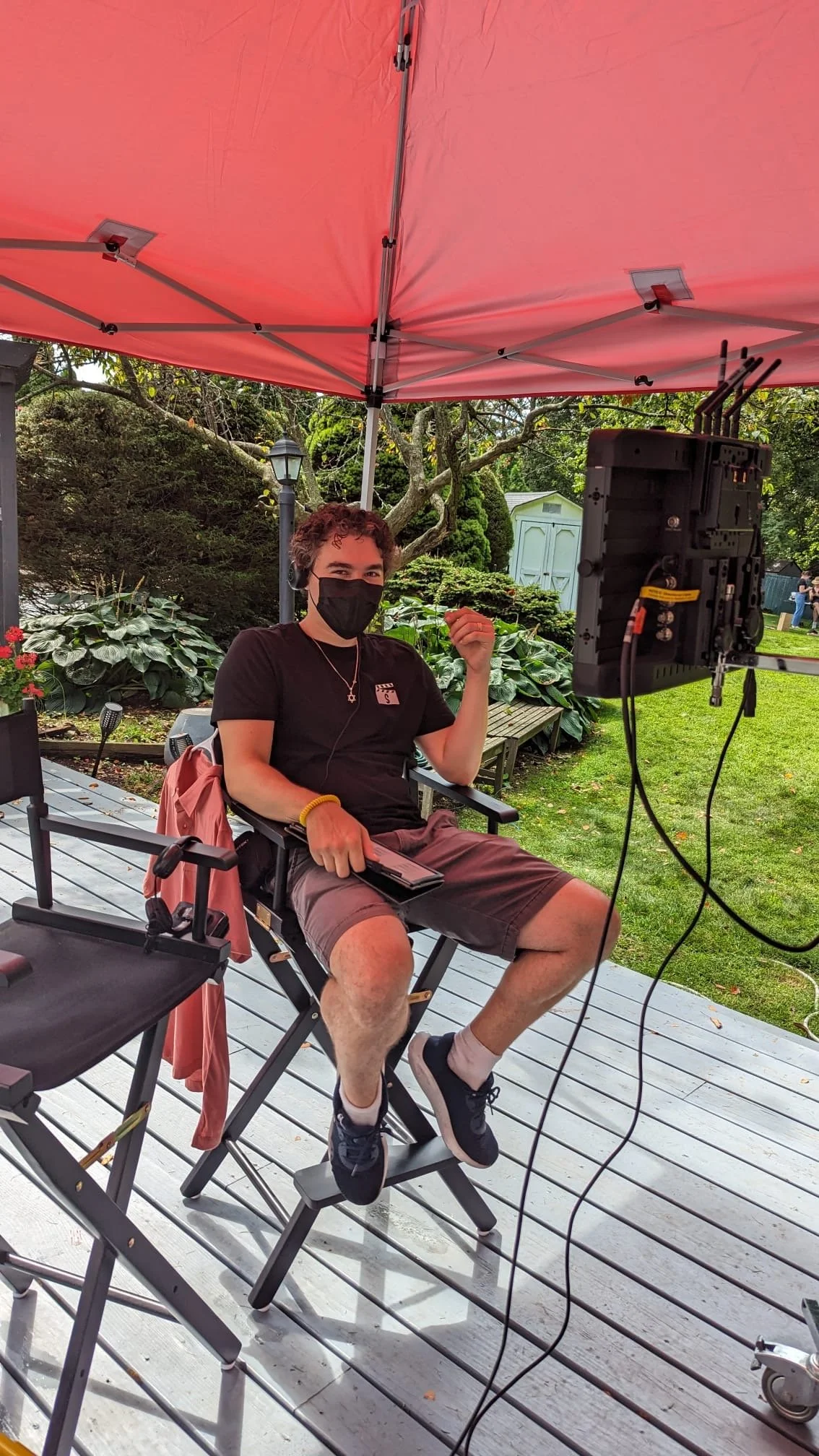Person sitting outdoors on director's chair, wearing a face mask, black t-shirt, shorts, and sneakers, under a pink canopy with filming equipment nearby.