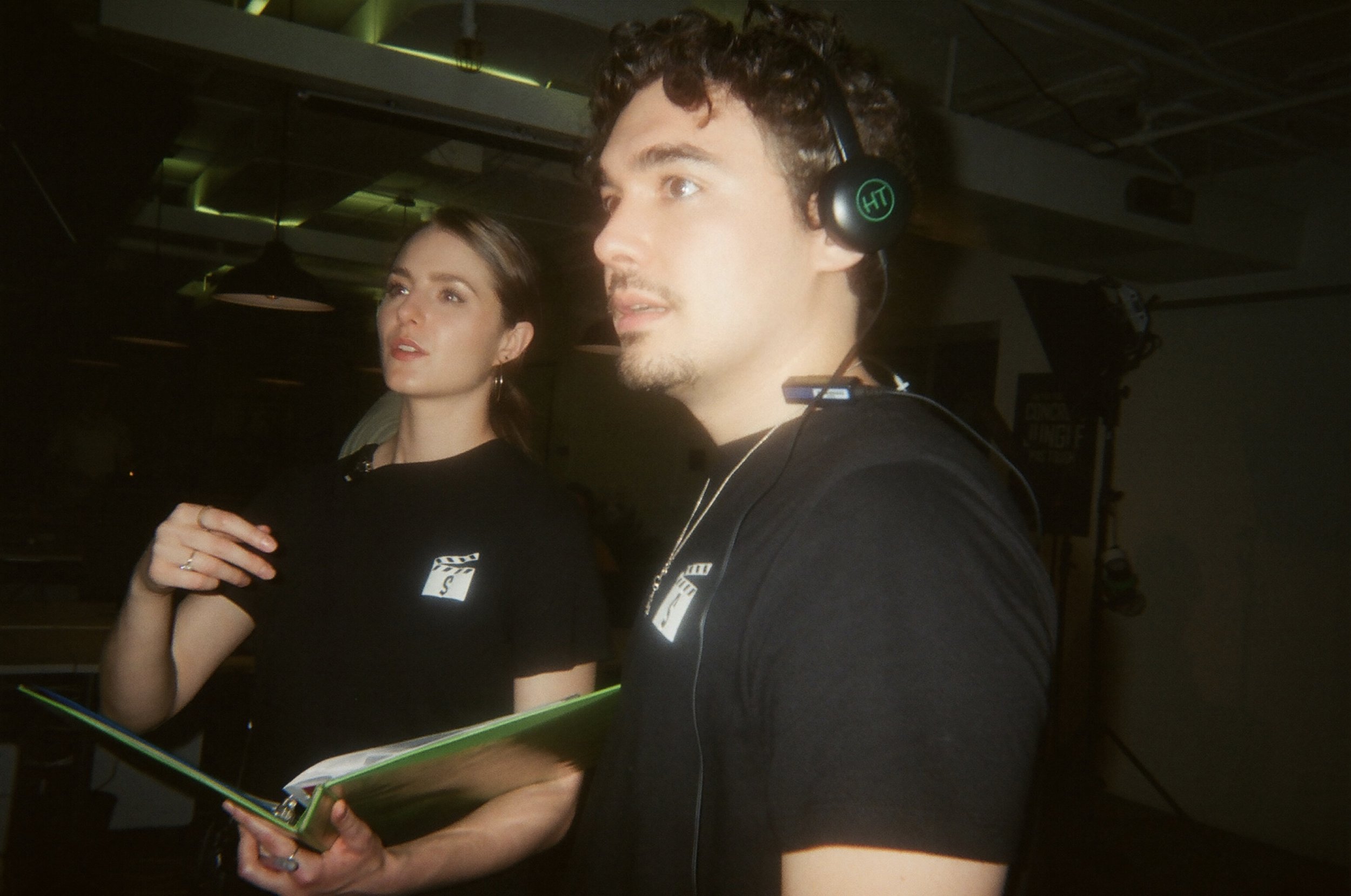Two young adults, a woman and a man, standing in a dimly lit room, possibly a studio or production setting. The woman has brown hair, wears hoop earrings, and holds a notebook. The man has curly hair, a goatee, and is wearing headphones.