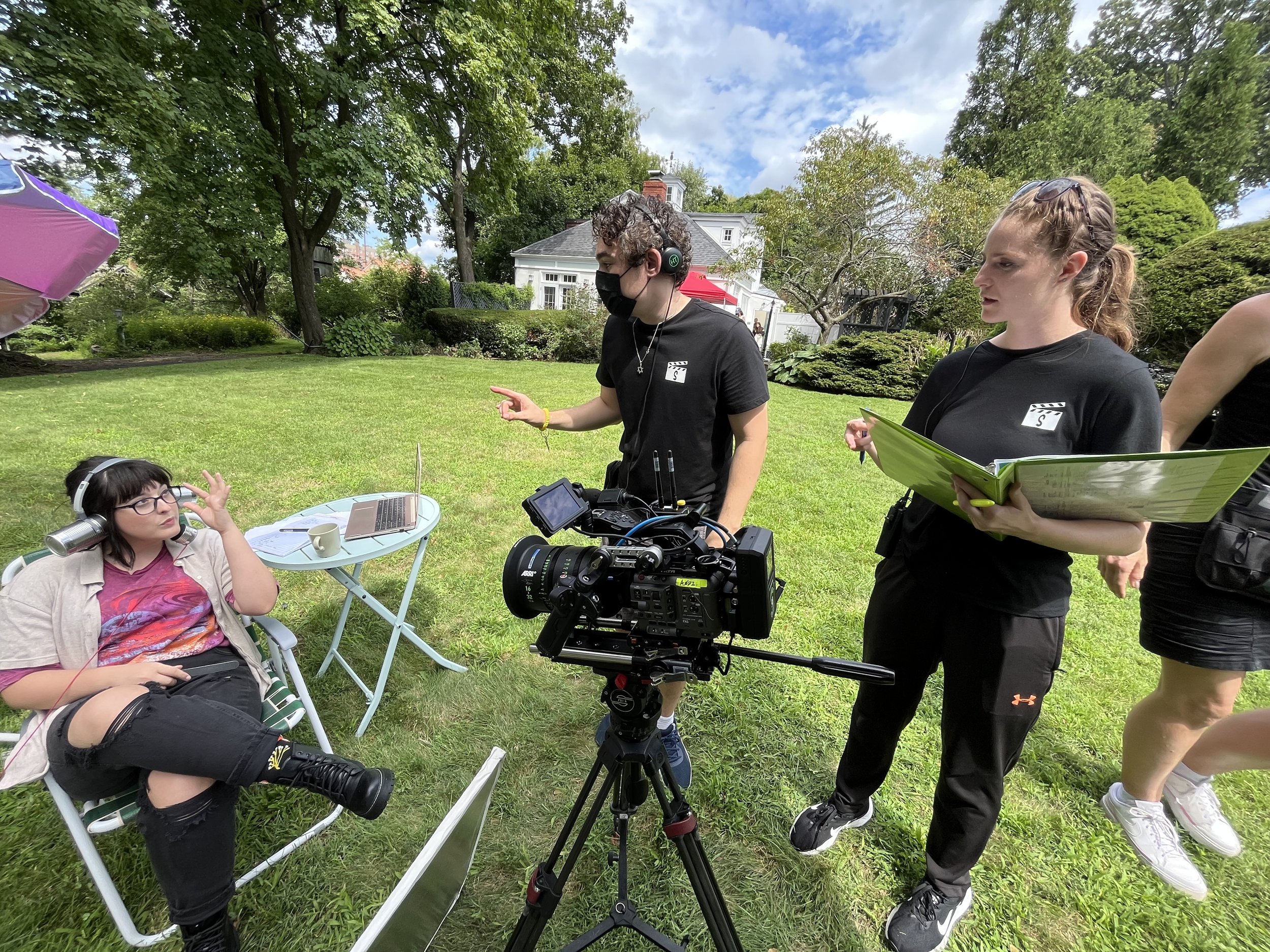 Filmmaker and crew conducting an outdoor interview with a young woman seated in a lawn chair on a sunny day with trees and a house in the background.