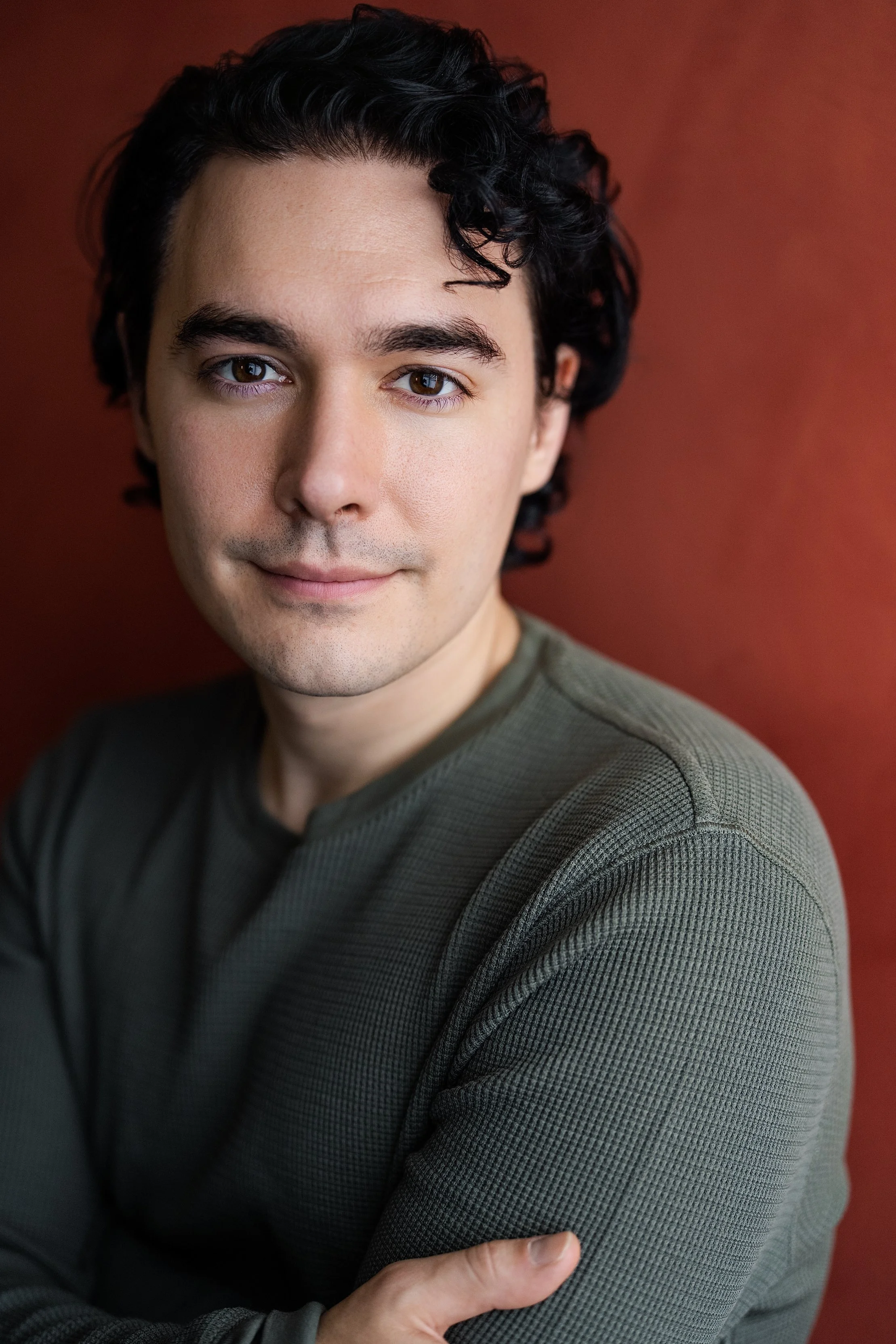 Portrait of a young man with dark, curly hair and light skin, wearing a green textured long-sleeve shirt, standing against a reddish-brown background.