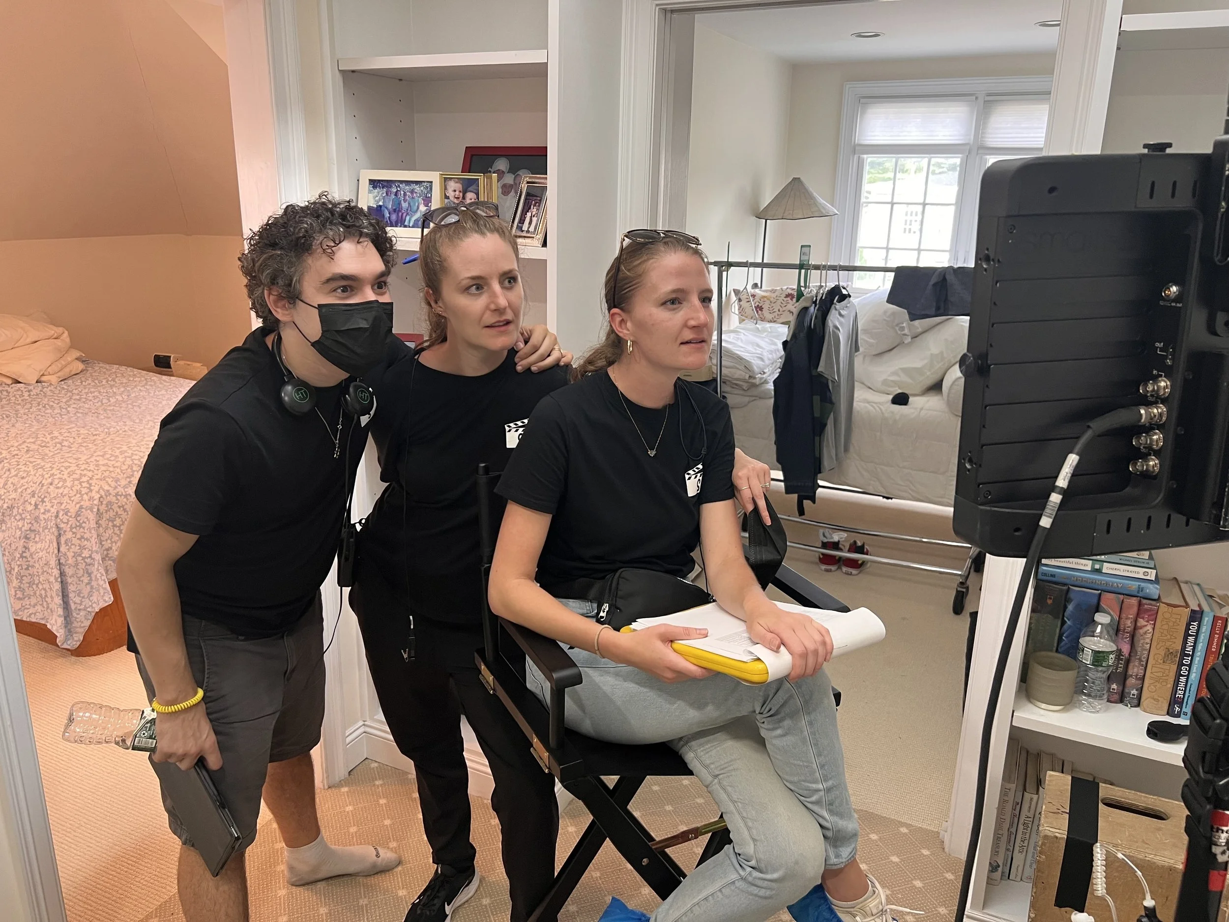 Three people on a film or video set. One woman is sitting on a director’s chair with a remote control or monitor, surrounded by two crew members, one of whom is wearing a face mask. They are focused on a screen or camera in front of them, with a bedr