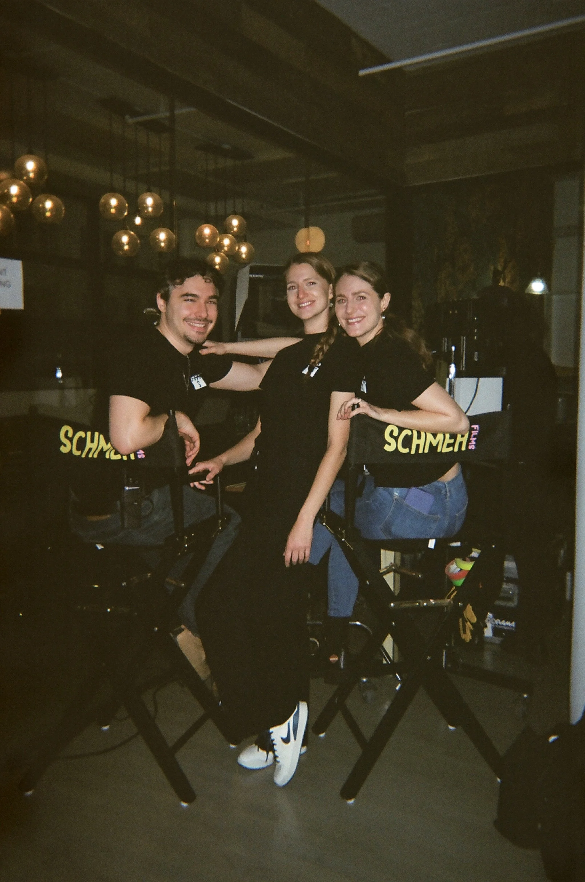 Three people sitting together on a stationary bicycle, smiling at the camera, inside a dimly lit room with modern decor, gold spherical light fixtures, and a background with a TV and other equipment.