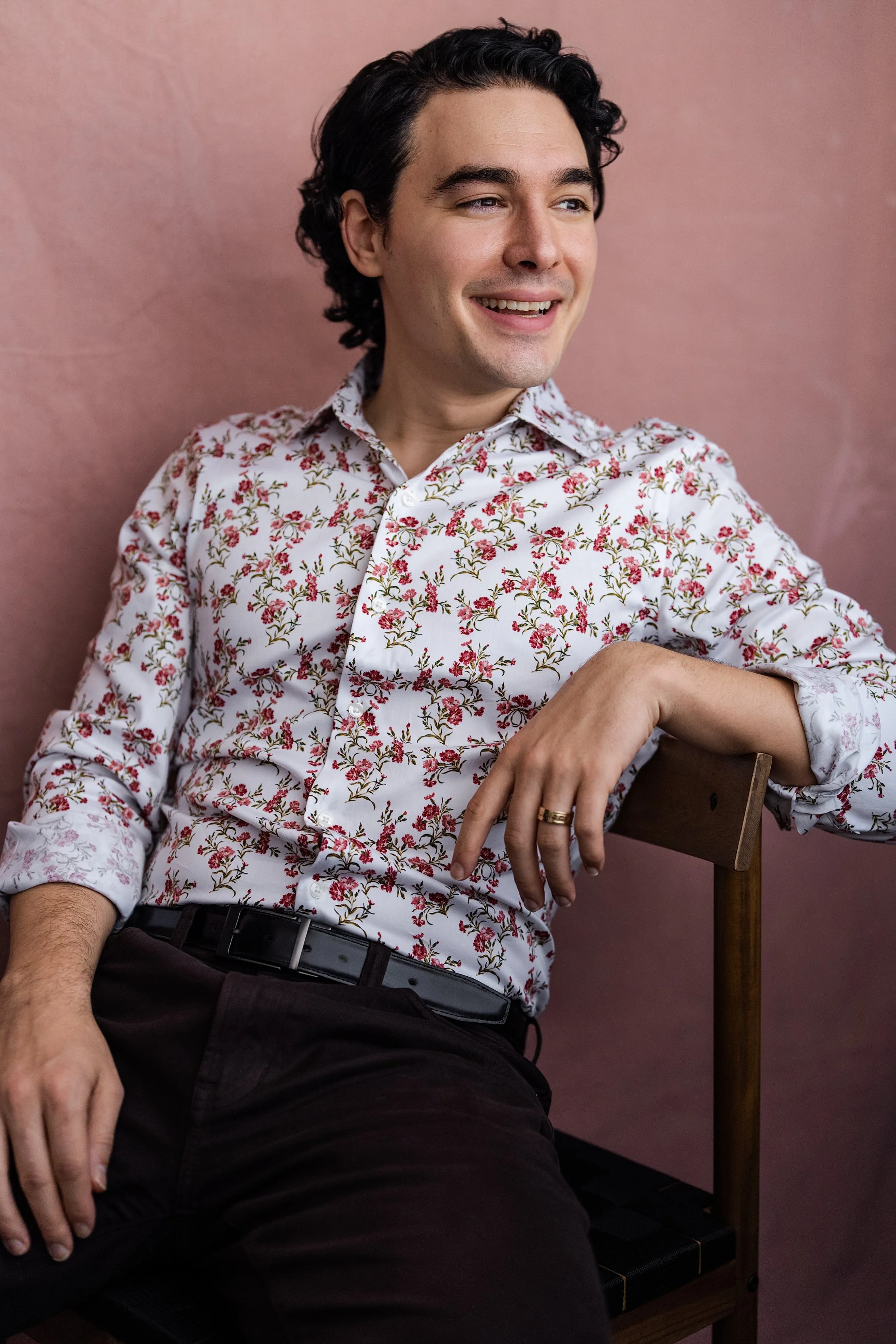 A young man with dark, curly hair sitting on a wooden chair against a pink wall, wearing a white shirt with a pink and green floral pattern, black pants, and a black belt, smiling and looking to the side.