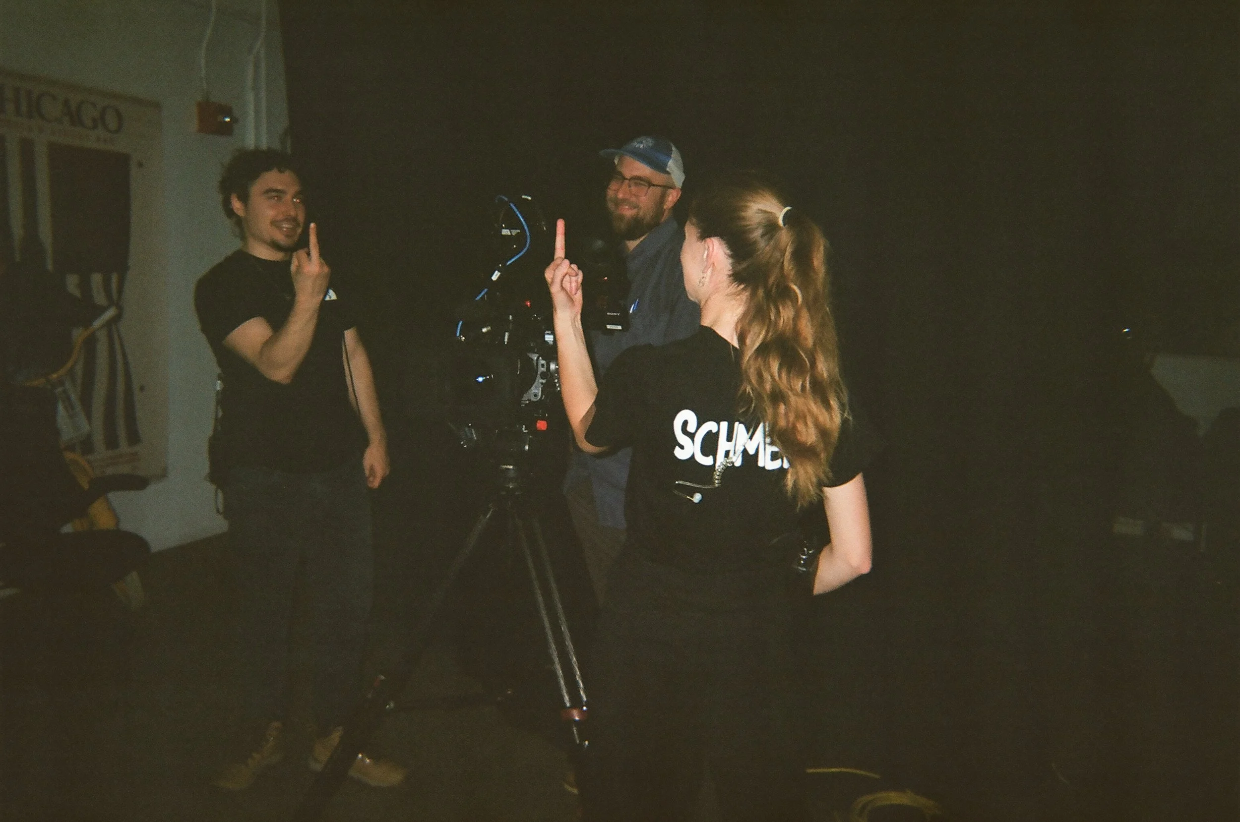 Three people in a dimly lit room, two of them wearing black shirts with one labeled 'SCHME'; they are engaged in filming or photography, with one person holding a camera and two others gesturing with one finger up.