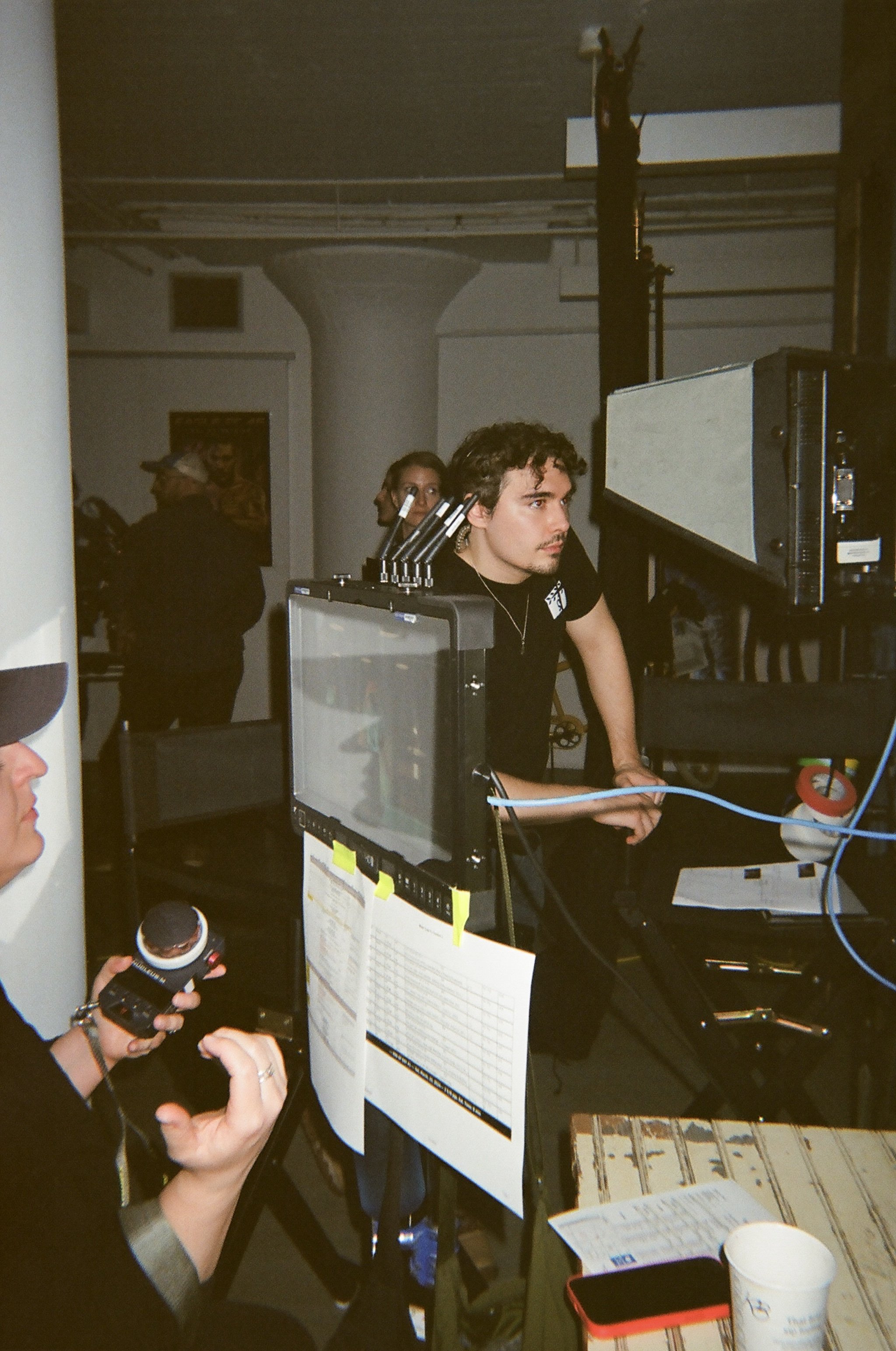 A man with dark hair and a mustache operating a camera on a film or TV set, with various equipment and crew members in the background.