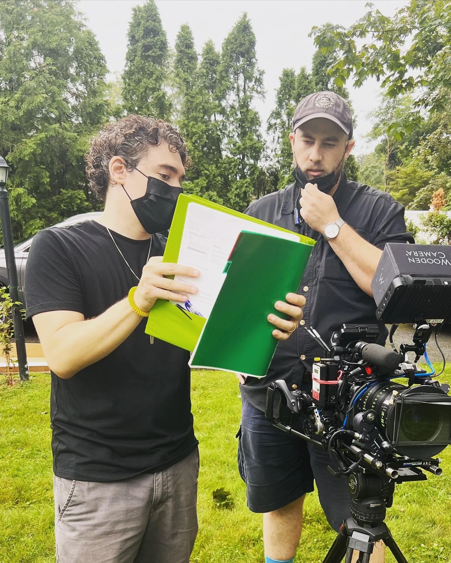 Two men wearing black face masks standing outdoors, looking at a clipboard with documents. One man is holding the clipboard, while the other is standing next to a professional film camera on a tripod. The background shows green trees and a lawn.