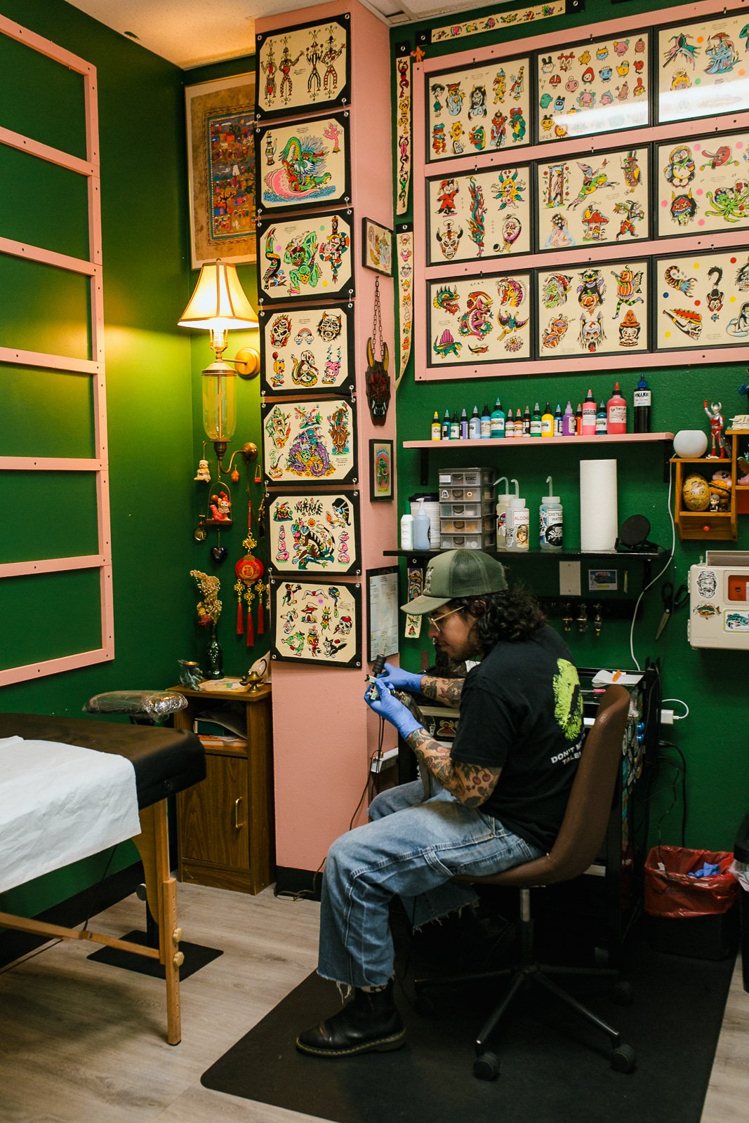 Tattoo artist working in a studio decorated with traditional-style tattoo flash art on the walls, with various inks and supplies on a shelf behind him.