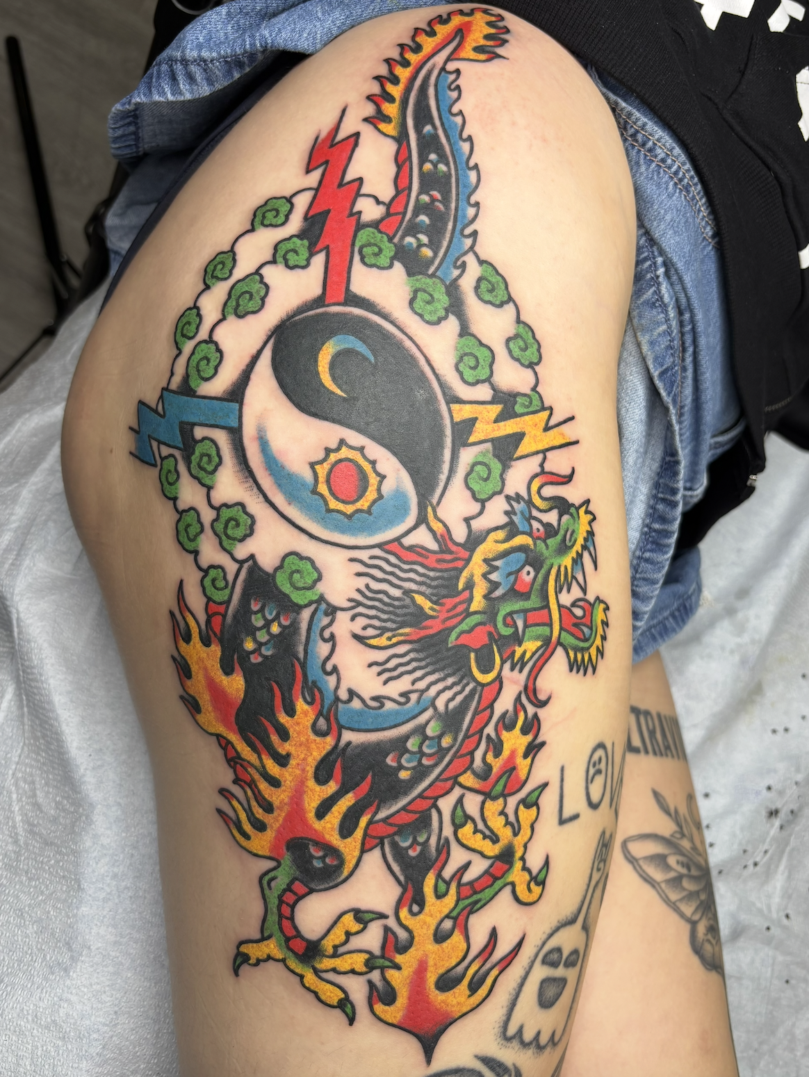 Colorful tattoo of a dragon intertwined with flames, lightning bolts, clouds, and a yin-yang symbol on a person's thigh.