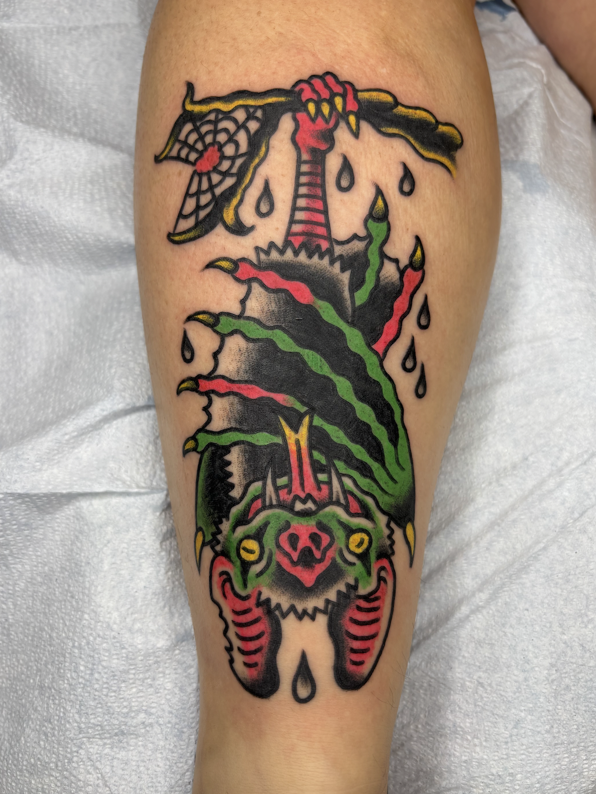 Tattoo of a dragon with a split face, featuring green and red colors, with flames and smoke, and a spider web with a red dot in the background.