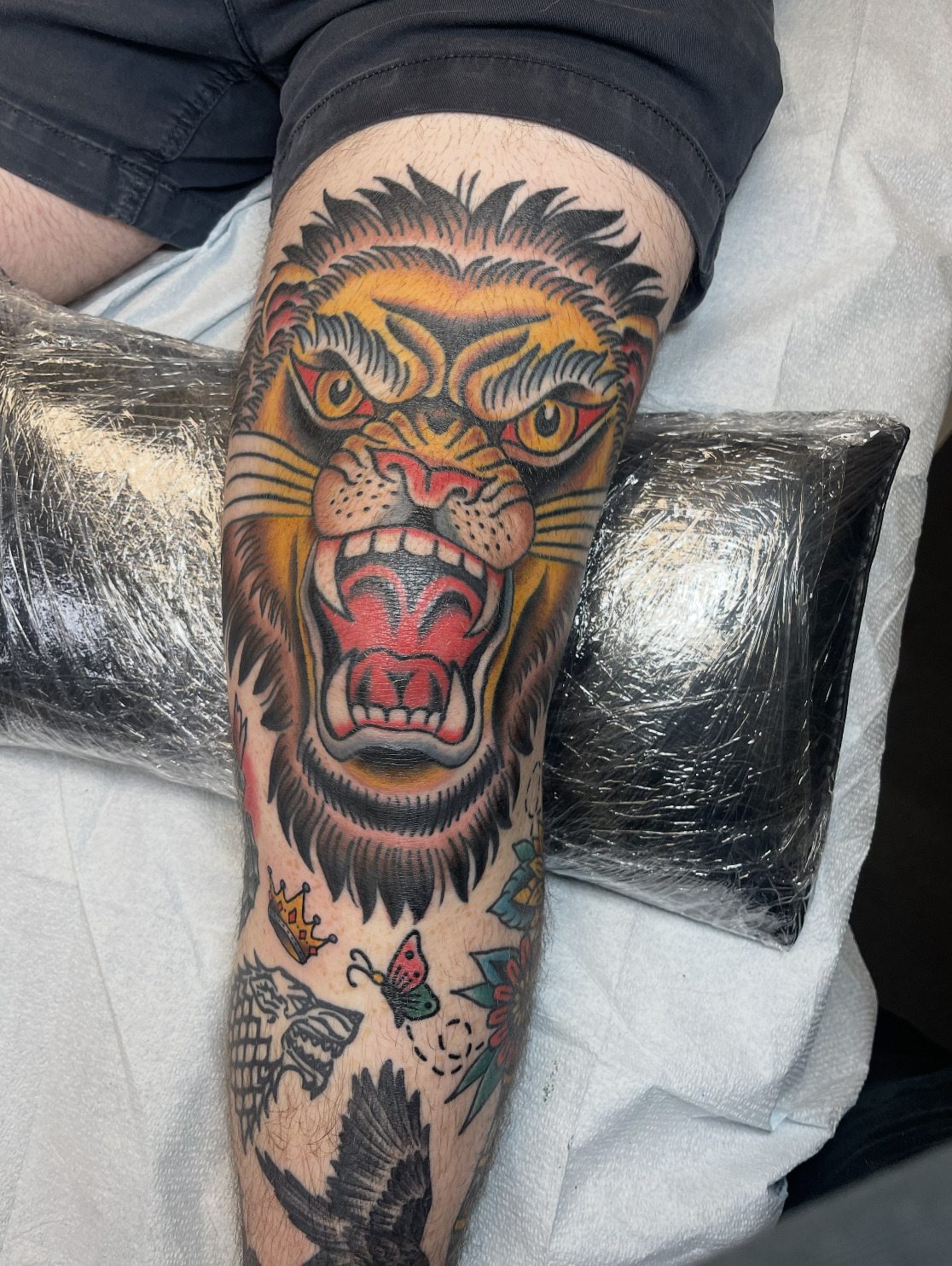 A colorful tattoo of a roaring lion's face on a person's forearm, with tattoos of a crown, butterfly, and bird below it.