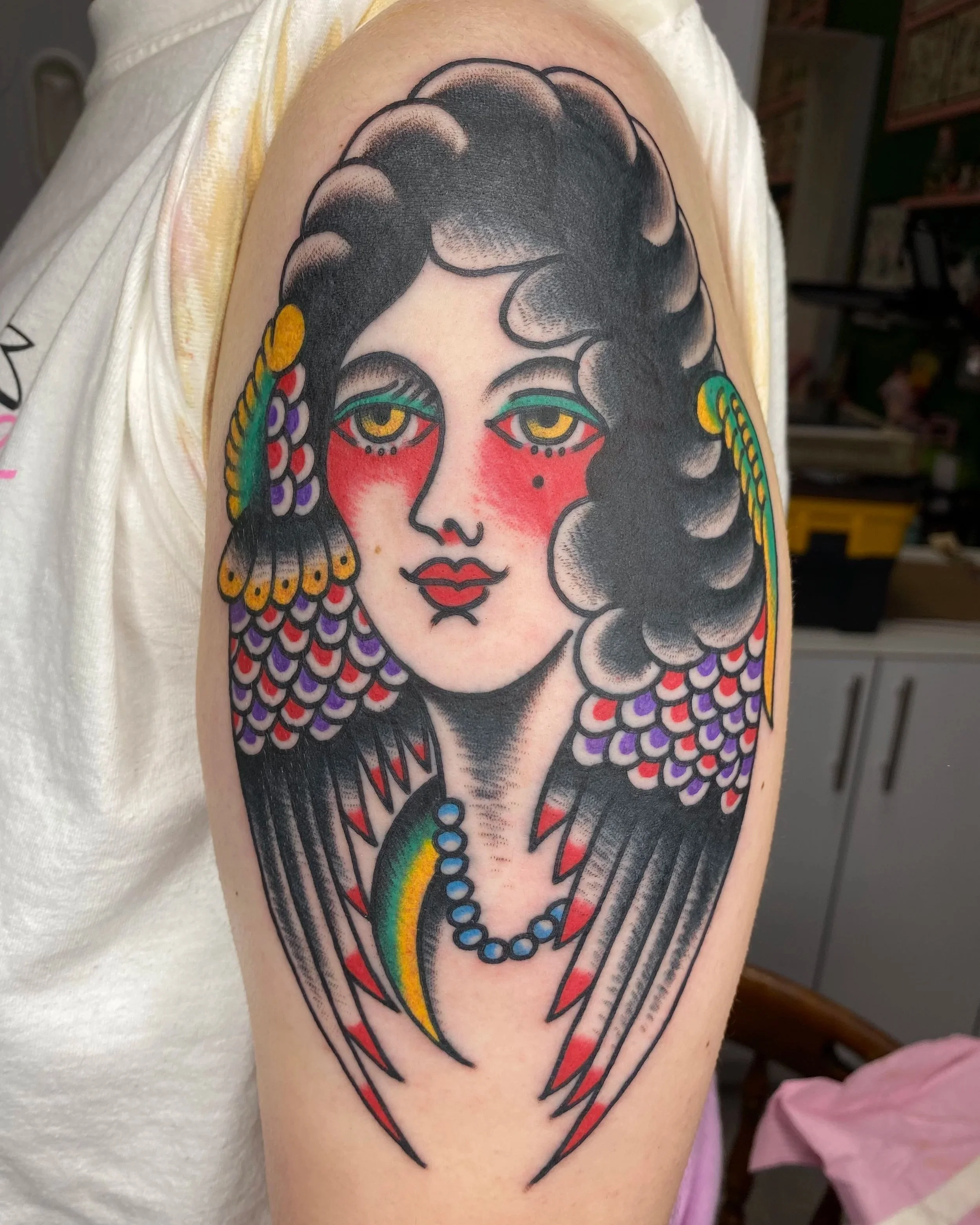 Tattoo of a stylized woman with black hair, yellow eyes, red cheeks, and rainbow-colored tears, wearing a beaded necklace and earrings.