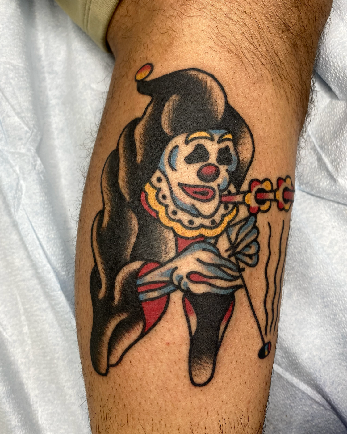 Tattoo of a clown with exaggerated facial features, holding a clown mask, with colorful details and black outlines.
