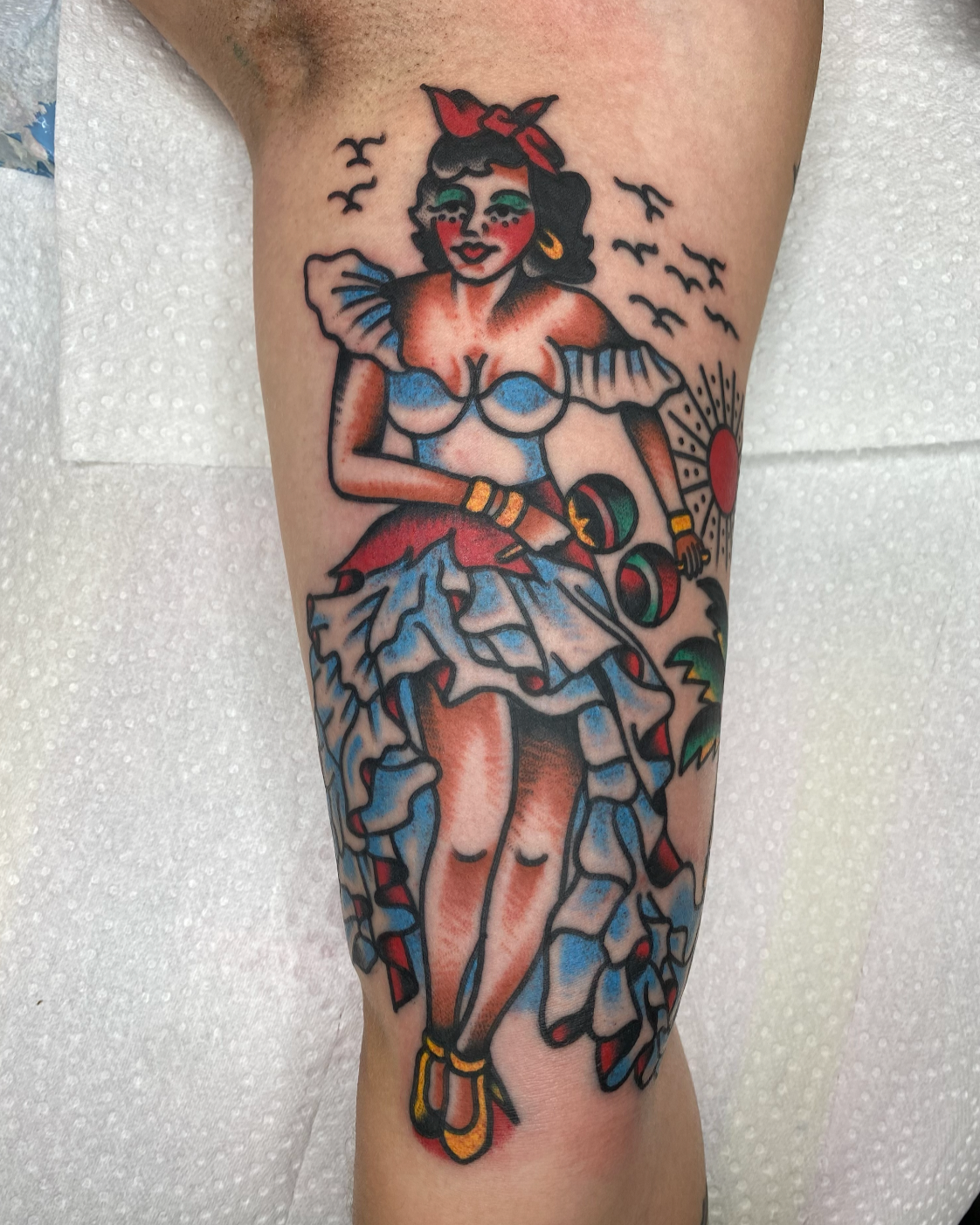 Tattoo of a pin-up style woman with dark hair, red bandana, and blue dress, holding maracas, surrounded by birds and a sun, with vibrant colors and bold outlines.