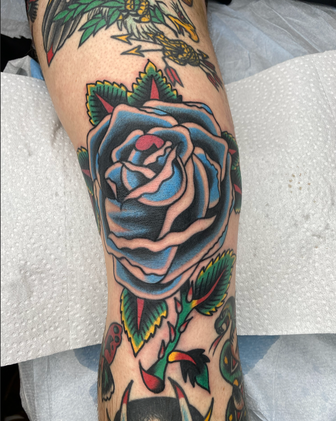 Colorful tattoo of a blue and black rose with green leaves and red accents on a person's leg.