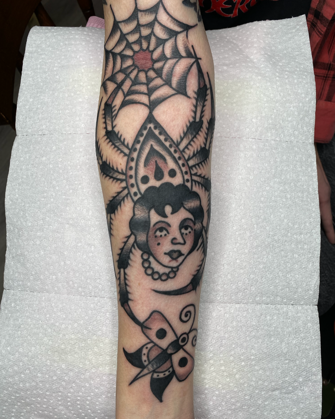 Tattoo on arm featuring a woman with short hair and a pearl necklace, spider web and spider design, and a moth with symmetrical patterns.
