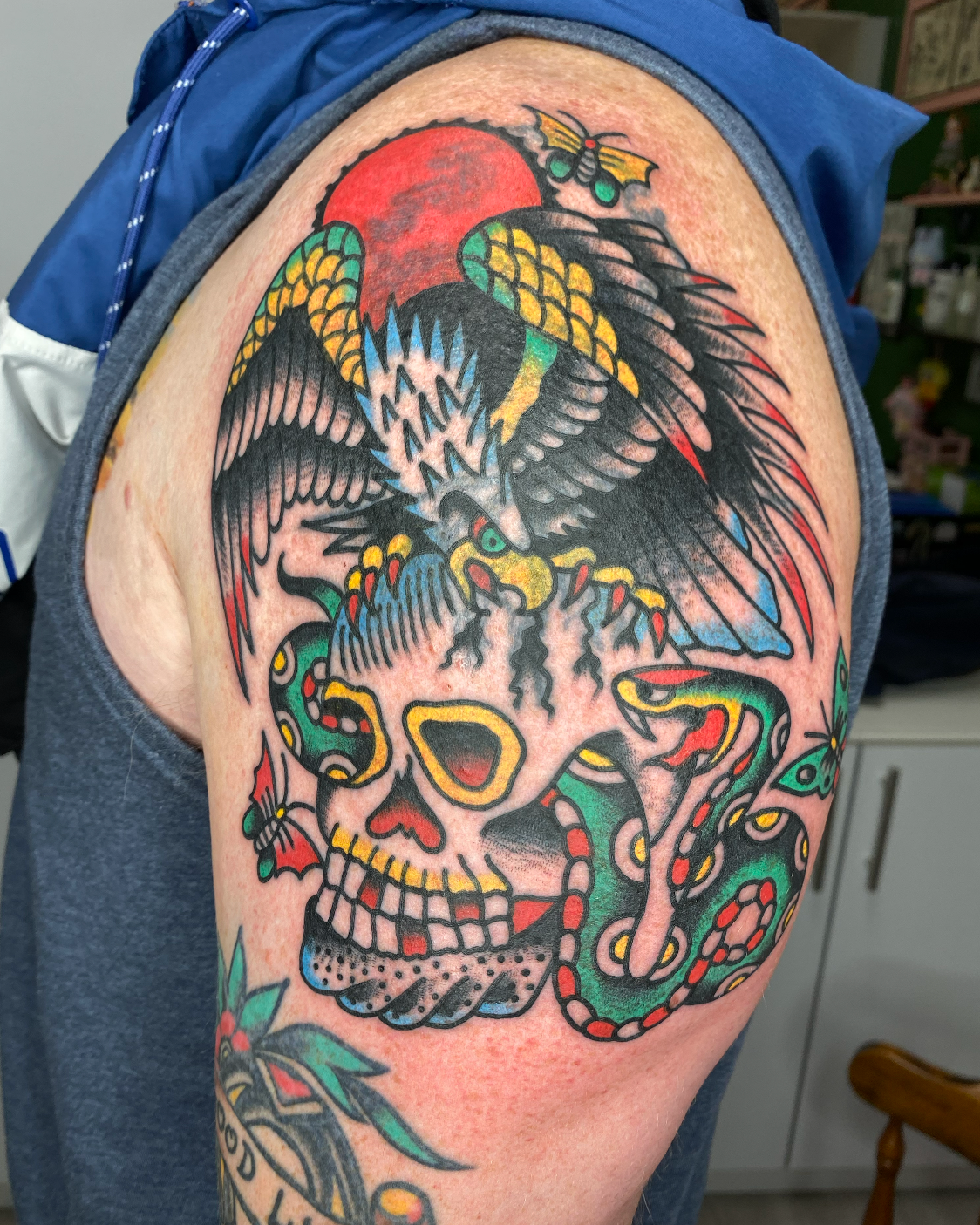 Tattoo of a stylized eagle with outstretched wings, perched above a skull, with a red sun and a moth in the background, all in bold, traditional tattoo style with vibrant colors.