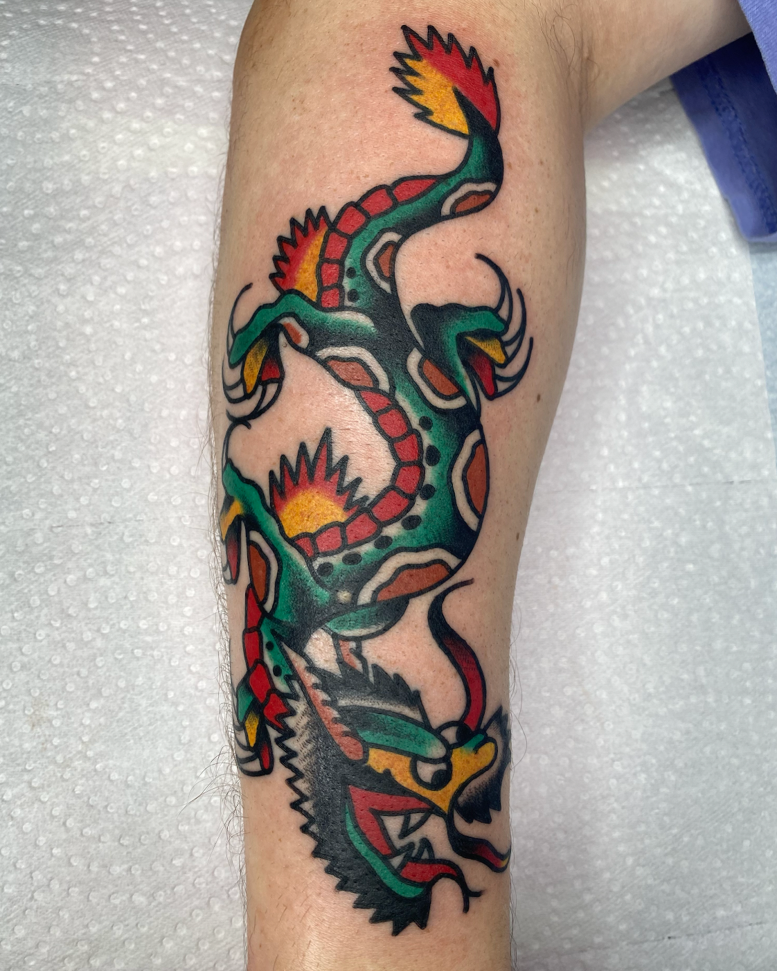 Colorful tattoo of a dragon with red, yellow, green, black, and orange details on a person's leg.