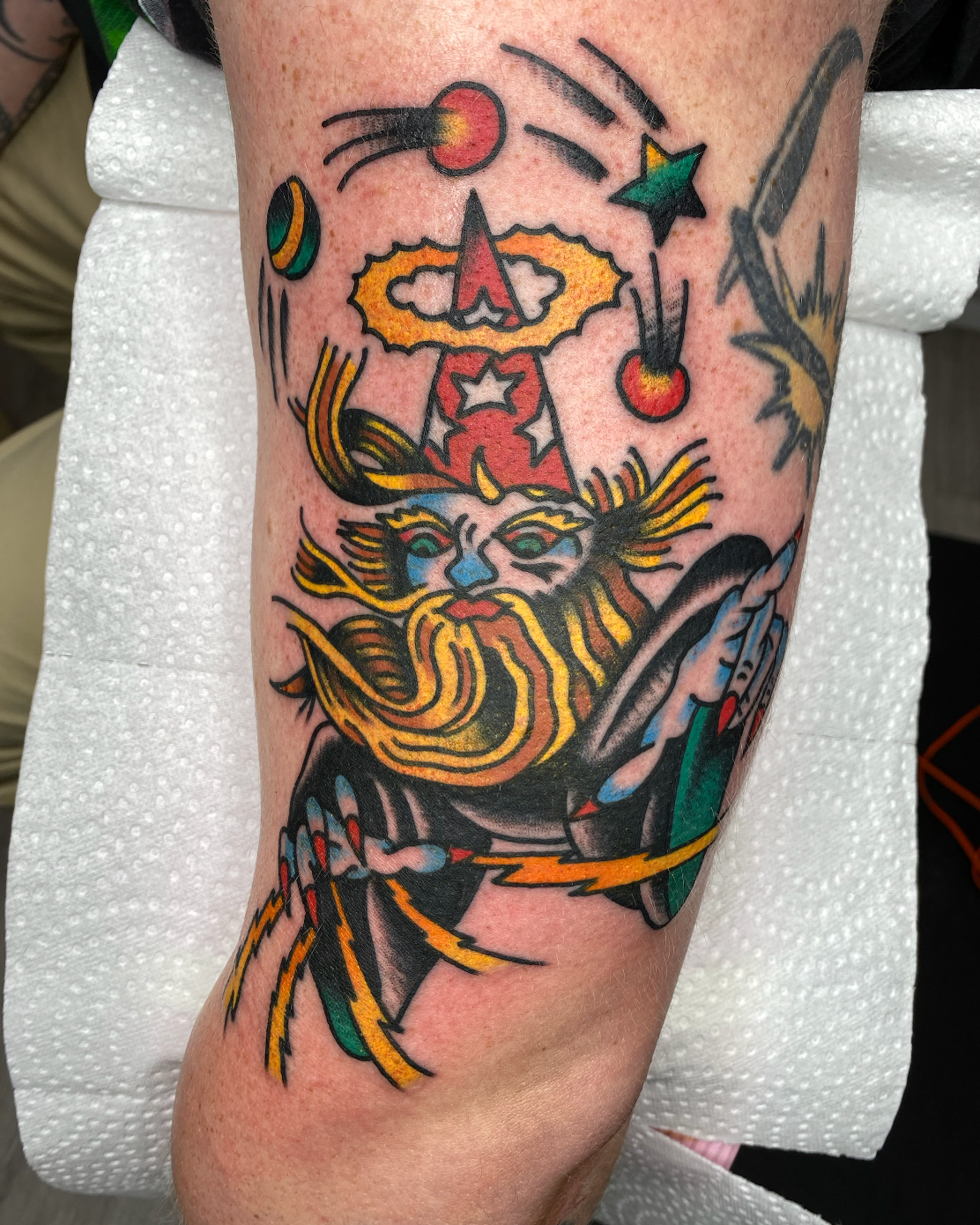 Colorful tattoo of a wizard with a long beard and flowing hair, holding a staff, with stars and comets around.
