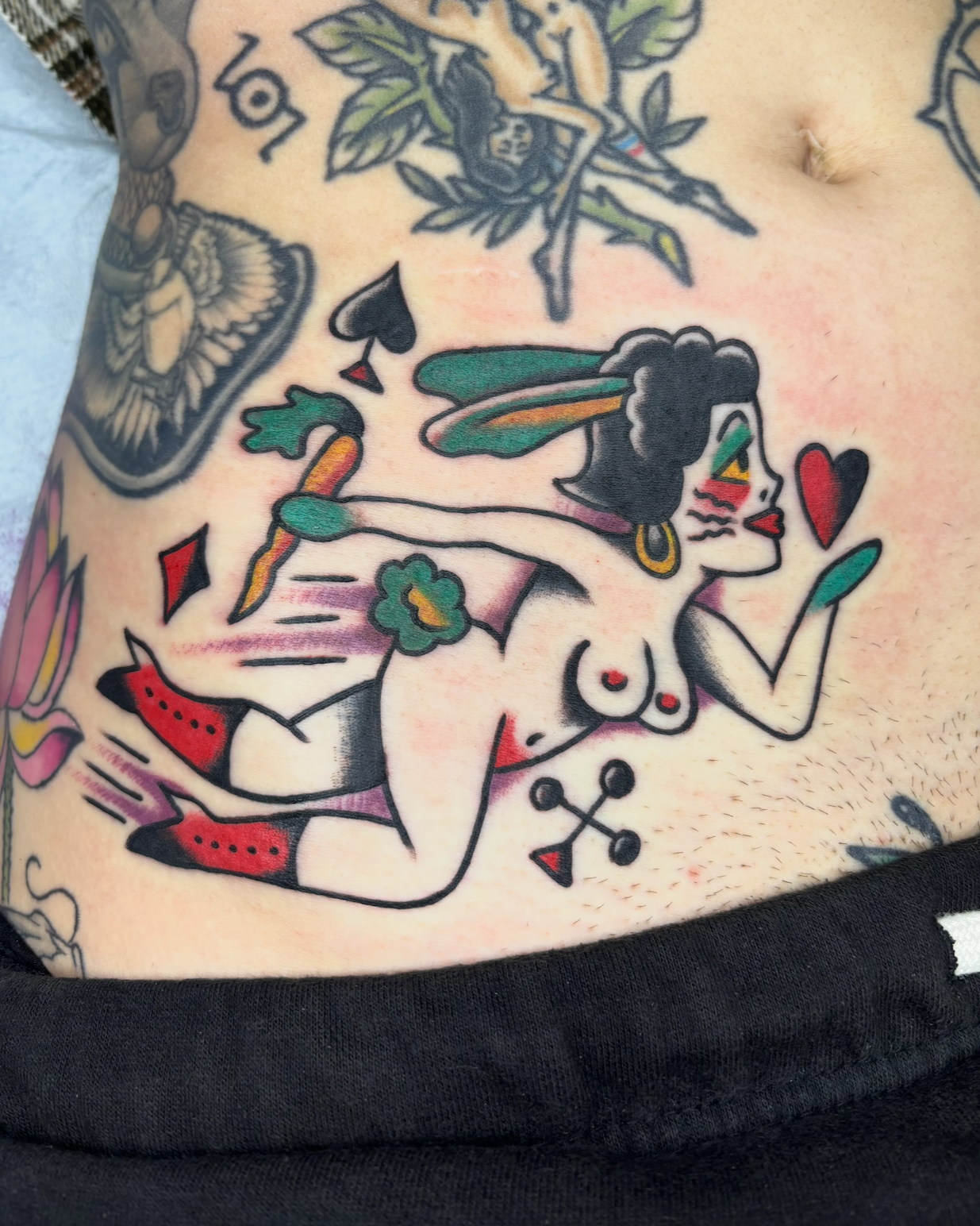 Tattoo of a woman with dark hair and green wings, wearing red boots and jewelry, holding a heart, with tattoos of a spider, a bird, and a rose.
