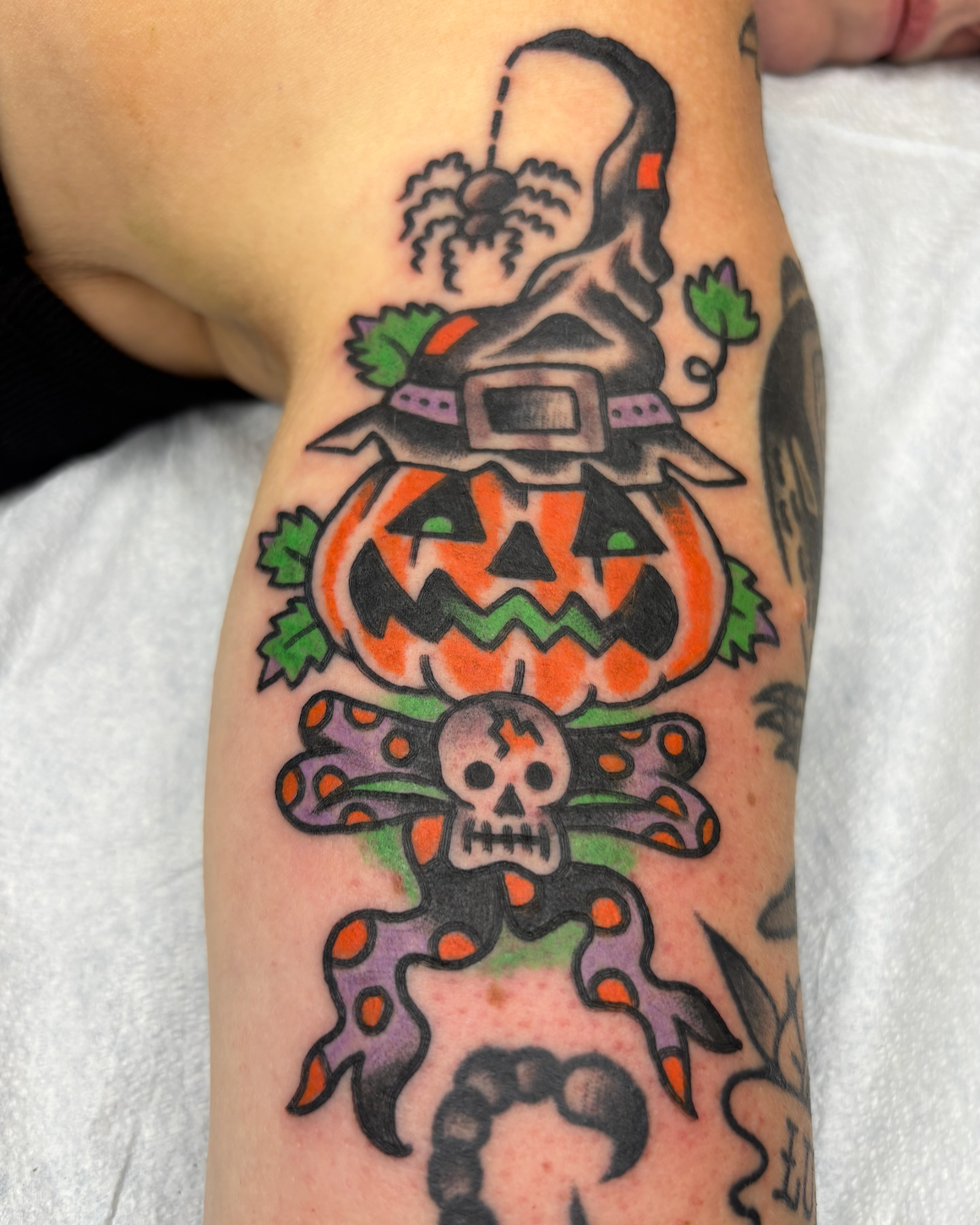 Tattoo of a carved pumpkin with a face, a skull, an octopus, a ghost, and green leaves on a person's forearm, celebrating Halloween.