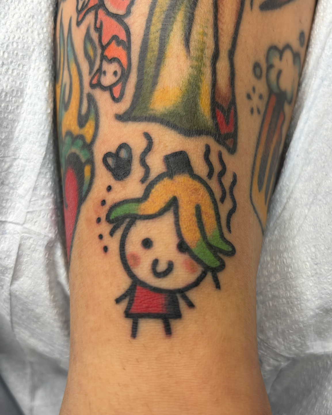 A colorful tattoo on a person's arm features a cartoon girl with a rainbow-colored mane of hair, a smiling face with blush cheeks, and a small red dress.