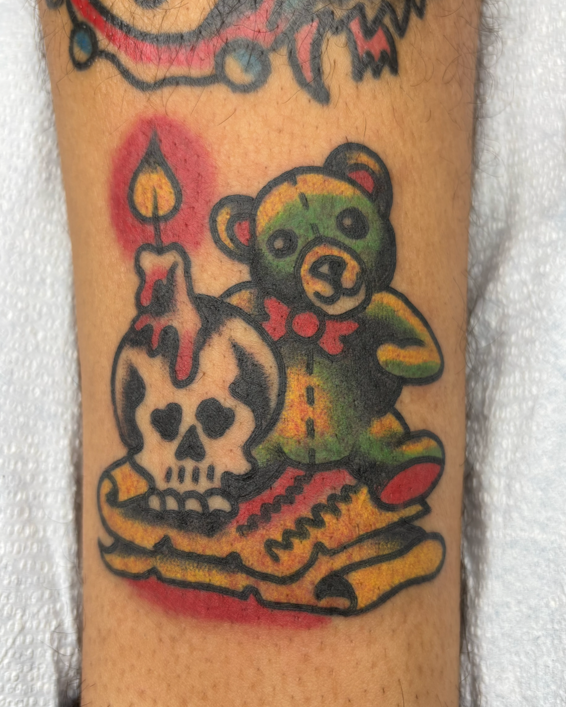 A colorful tattoo of a teddy bear holding a skull with a candle on top. The teddy bear has a red bow tie and the skull has black eye sockets and nose hole. The tattoo is outlined in black and features shades of green, yellow, and red.