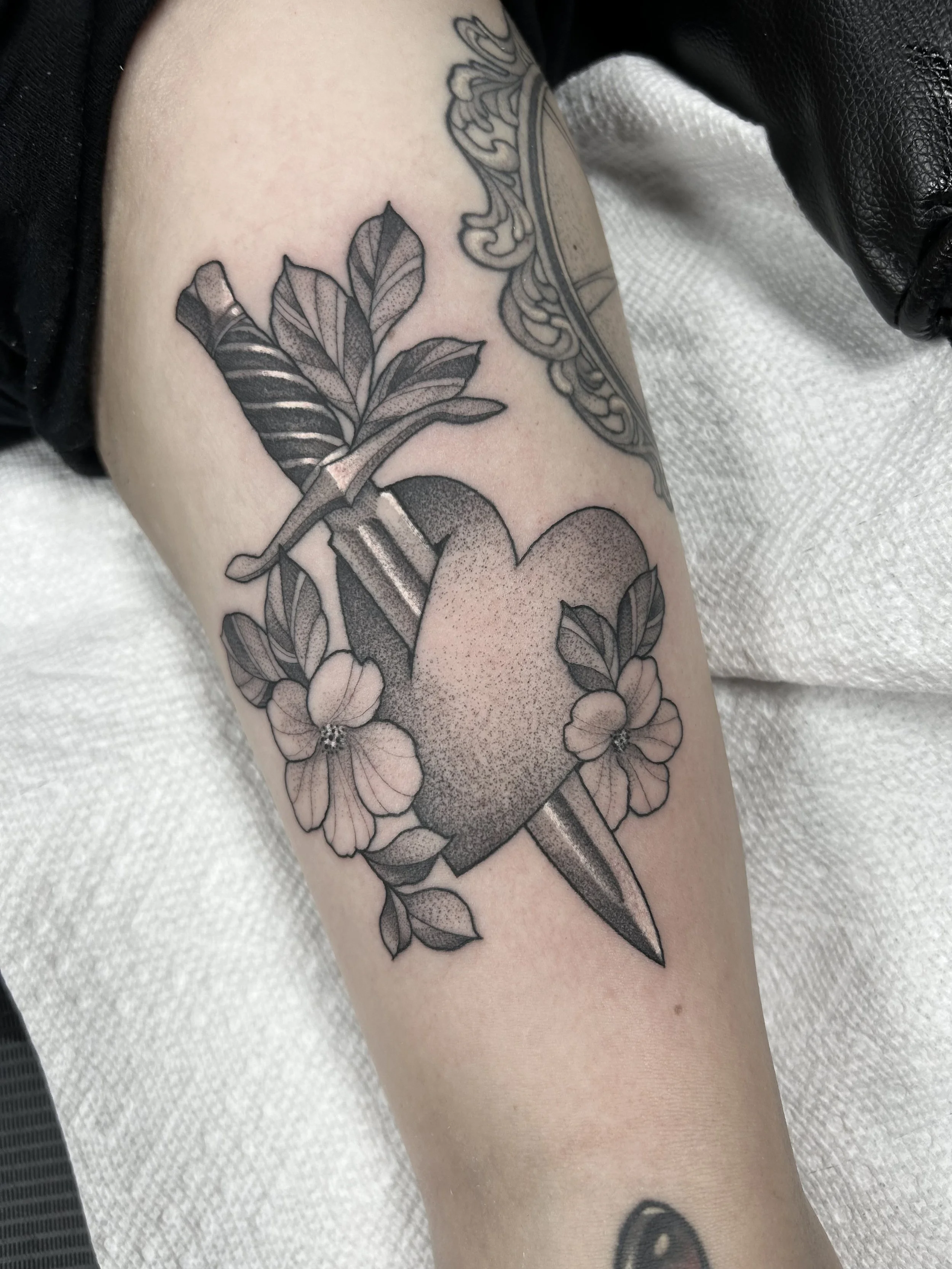 Black and gray tattoo of a heart with a dagger through it, surrounded by flowers and leaves on a person's arm.