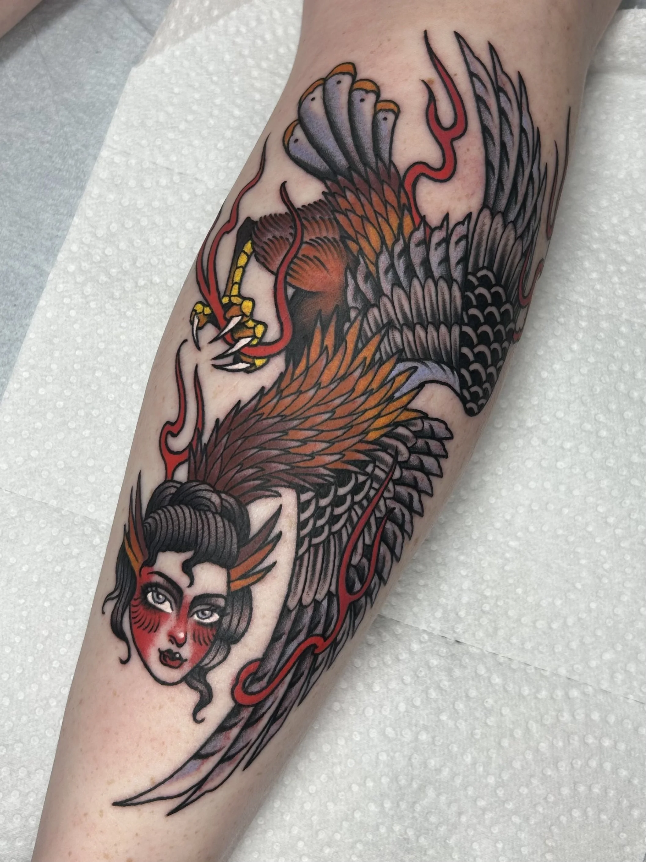 A tattoo of a woman with blue eyes and red face paint, with black and gray wings and red flames on her face, covering a person's arm.