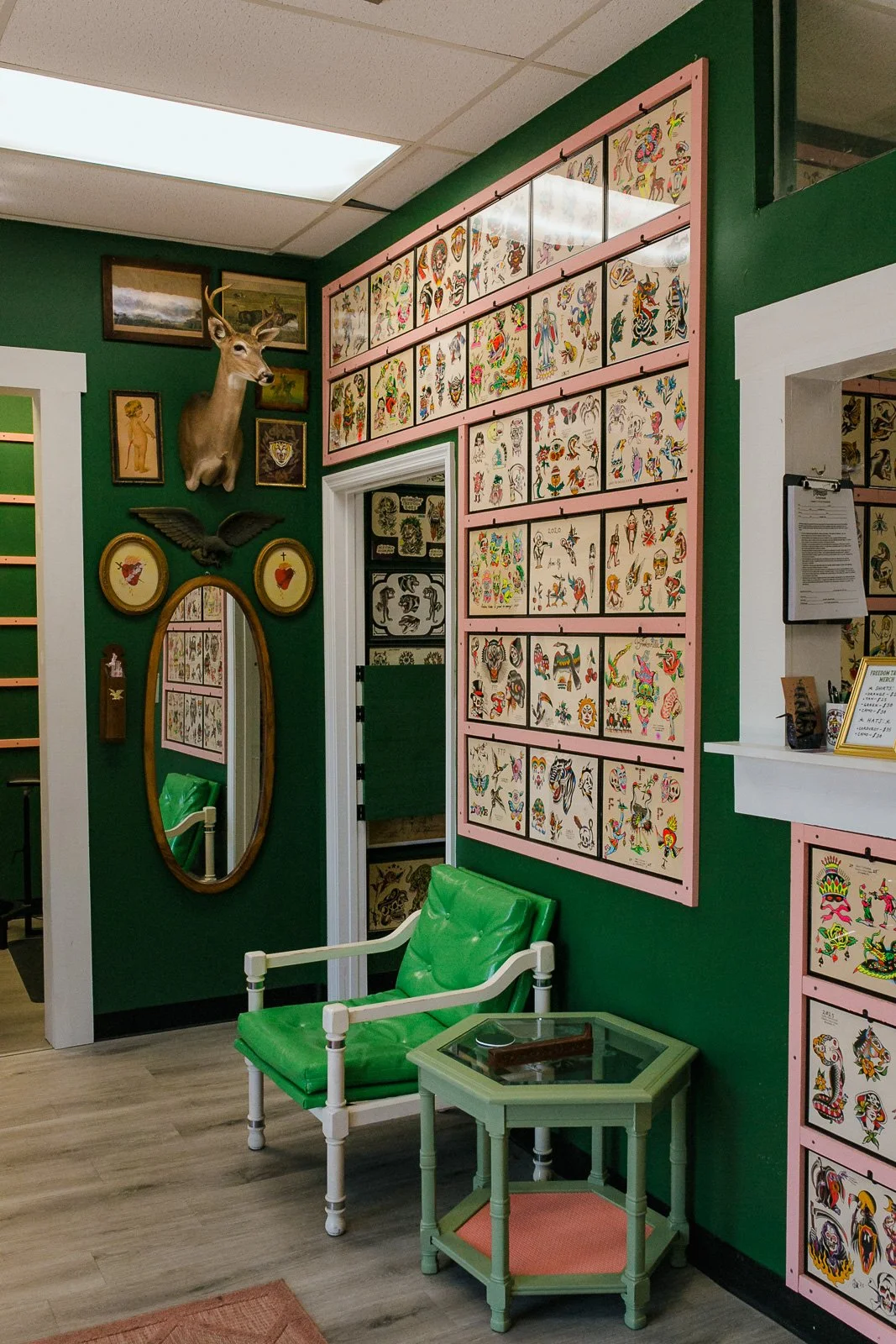 Interior of a tattoo shop with a green wall, featuring a mounted deer head, framed artwork, a large display of tattoo designs on pink-bordered panels, a green armchair, a small green side table with a glass top, and decorative framed art.