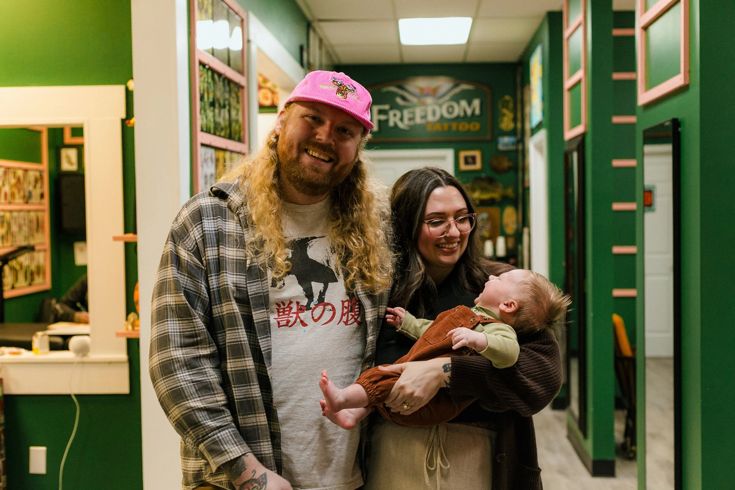 Two adults and a baby in a tattoo shop. The man has long blonde hair, tattoos, and wears a pink cap and a plaid shirt. The woman has dark hair, glasses, and holds the baby. The baby is lying on her arm and looks up at the woman. The background shows 